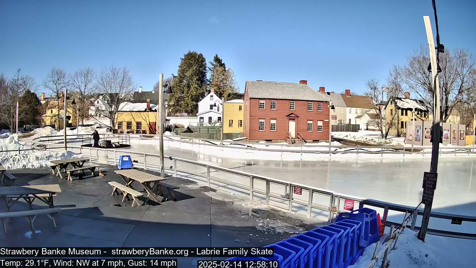 An outdoor ice skating rink is partially surrounded by historic-looking buildings on a sunny day with a northwest wind.