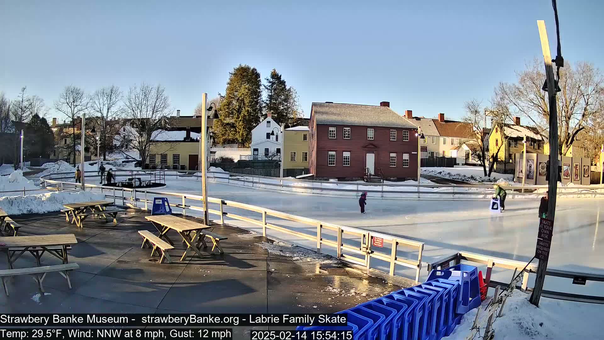 A sunny outdoor ice skating rink with several people skating, surrounded by historical buildings and picnic tables, in a snowy setting.