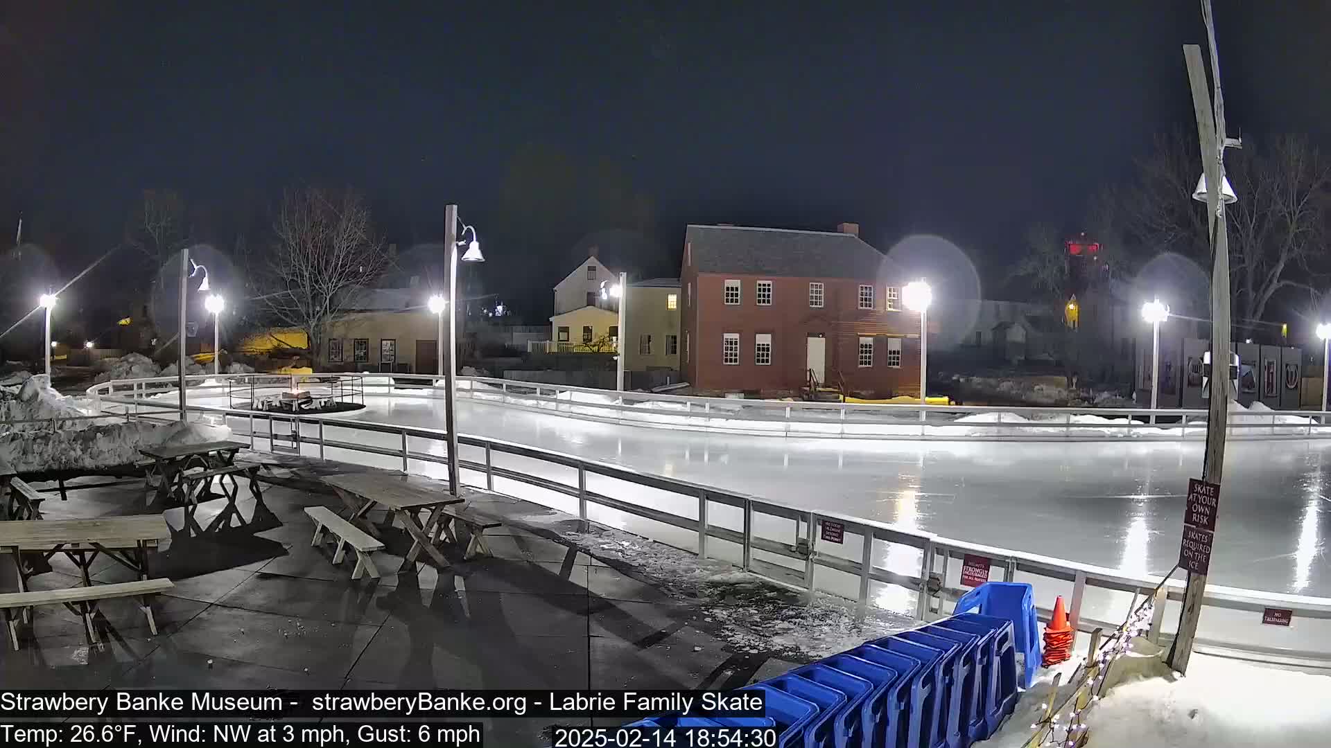 An outdoor ice rink is illuminated at night under partly cloudy skies, with picnic tables and blue plastic containers nearby.