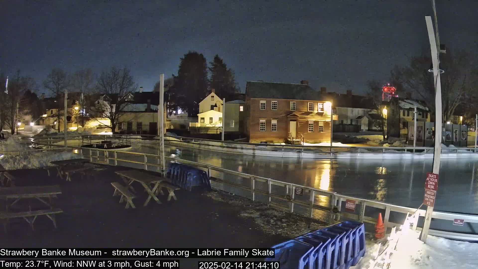A nighttime view of an outdoor ice skating rink, partially illuminated, next to several buildings under a dark, clear sky.