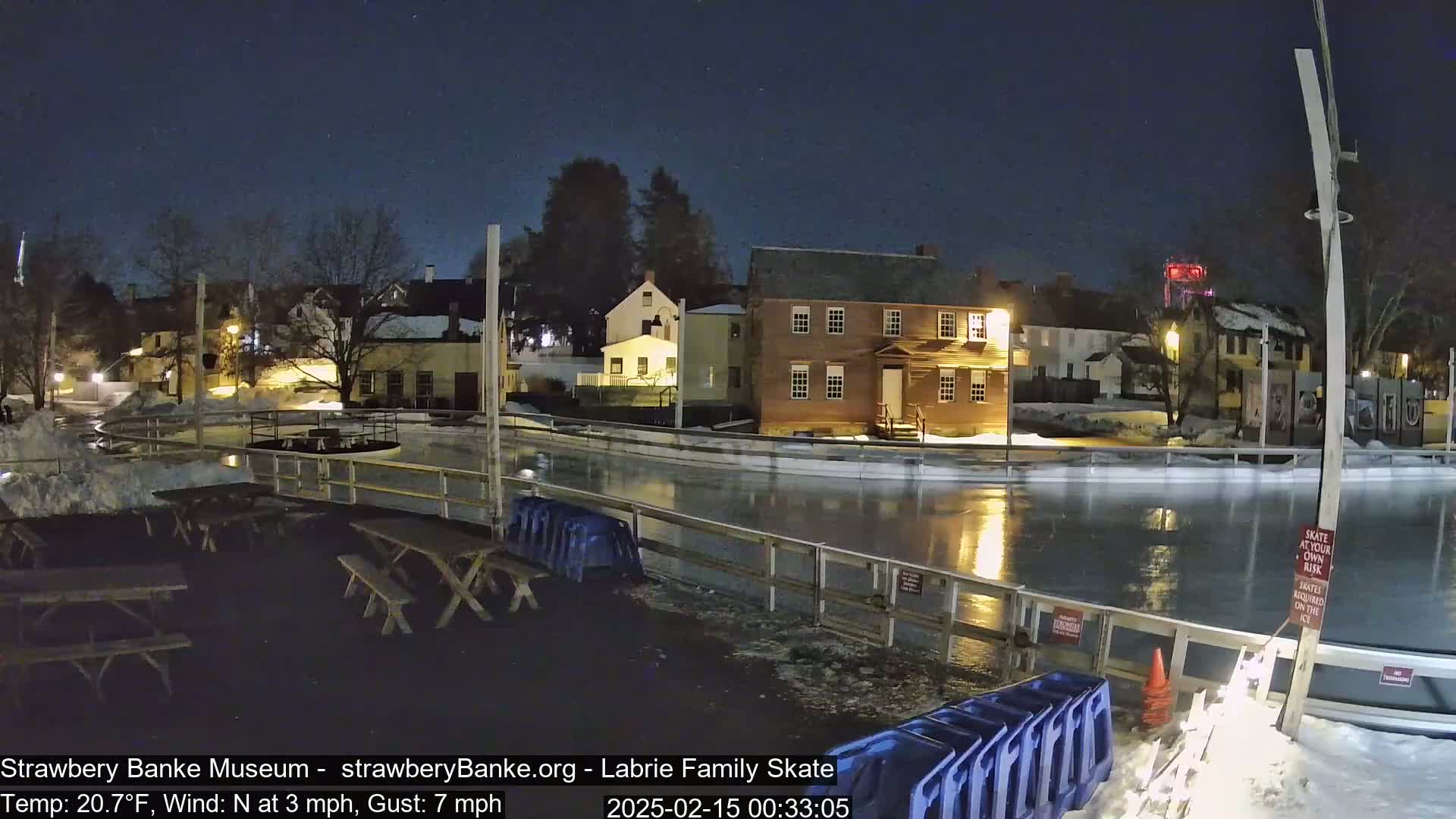 A nighttime view of an outdoor ice rink near several illuminated buildings under a clear, starry sky.