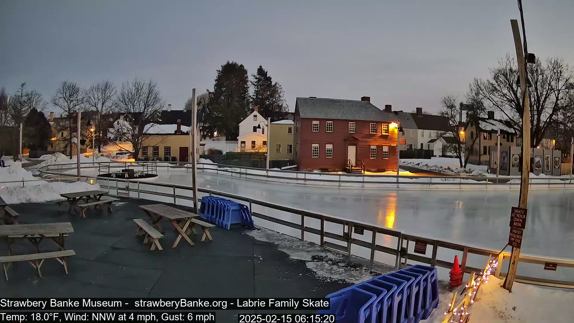 An outdoor ice skating rink is illuminated at dusk, surrounded by snow-covered ground and historic-looking buildings.