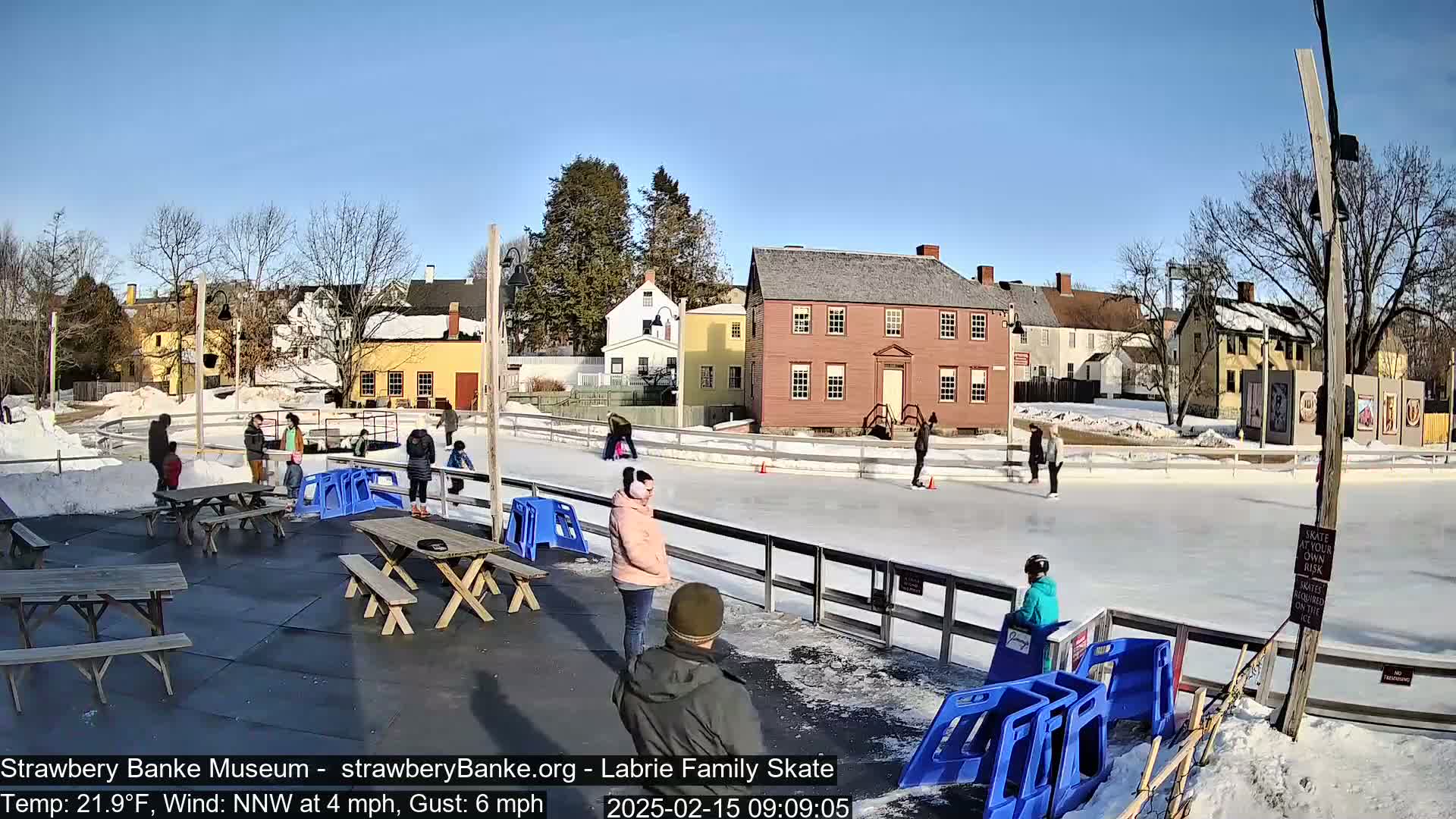 An outdoor ice skating rink, surrounded by historical buildings and picnic tables, on a sunny day with a light north-northwest wind.