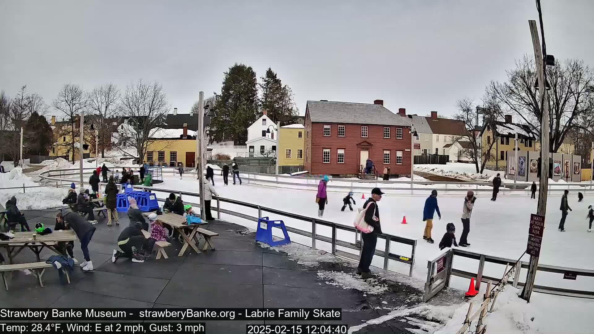 An outdoor ice skating rink, surrounded by historic buildings and people skating and watching, on a cloudy day with a temperature of 28.4°F and an east wind.