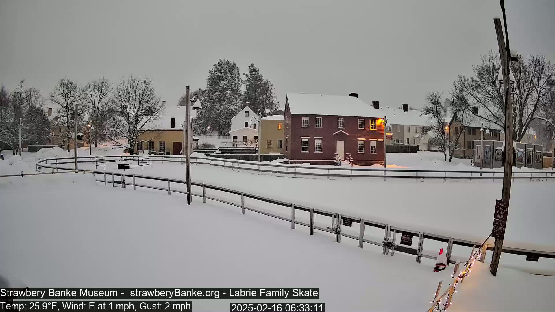A snow-covered outdoor skating rink is surrounded by historic-looking buildings under a gray sky during a light snowfall.