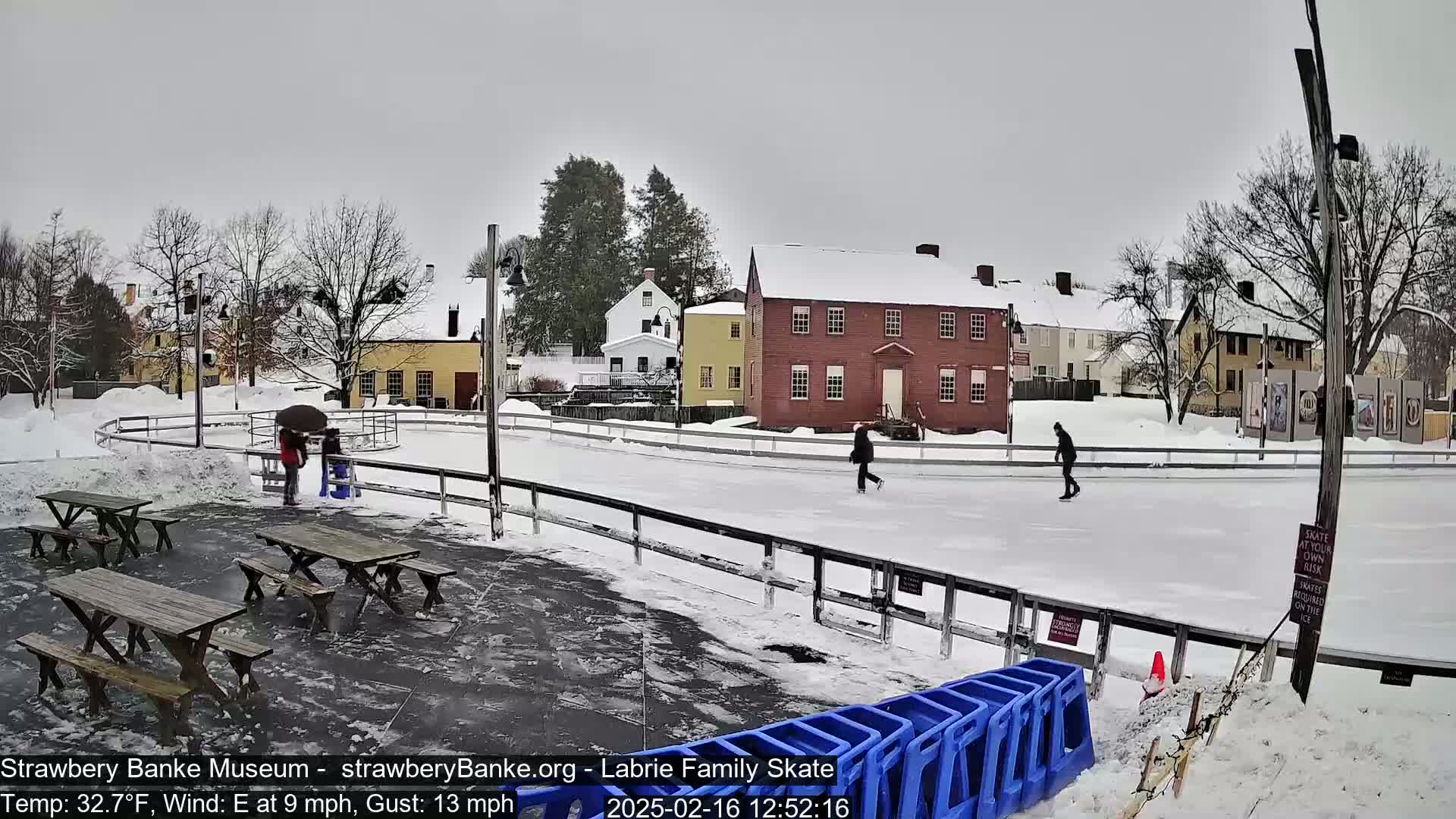An outdoor ice rink surrounded by snow-covered buildings and picnic tables under an overcast sky, with several people ice skating and others standing near the rink.
