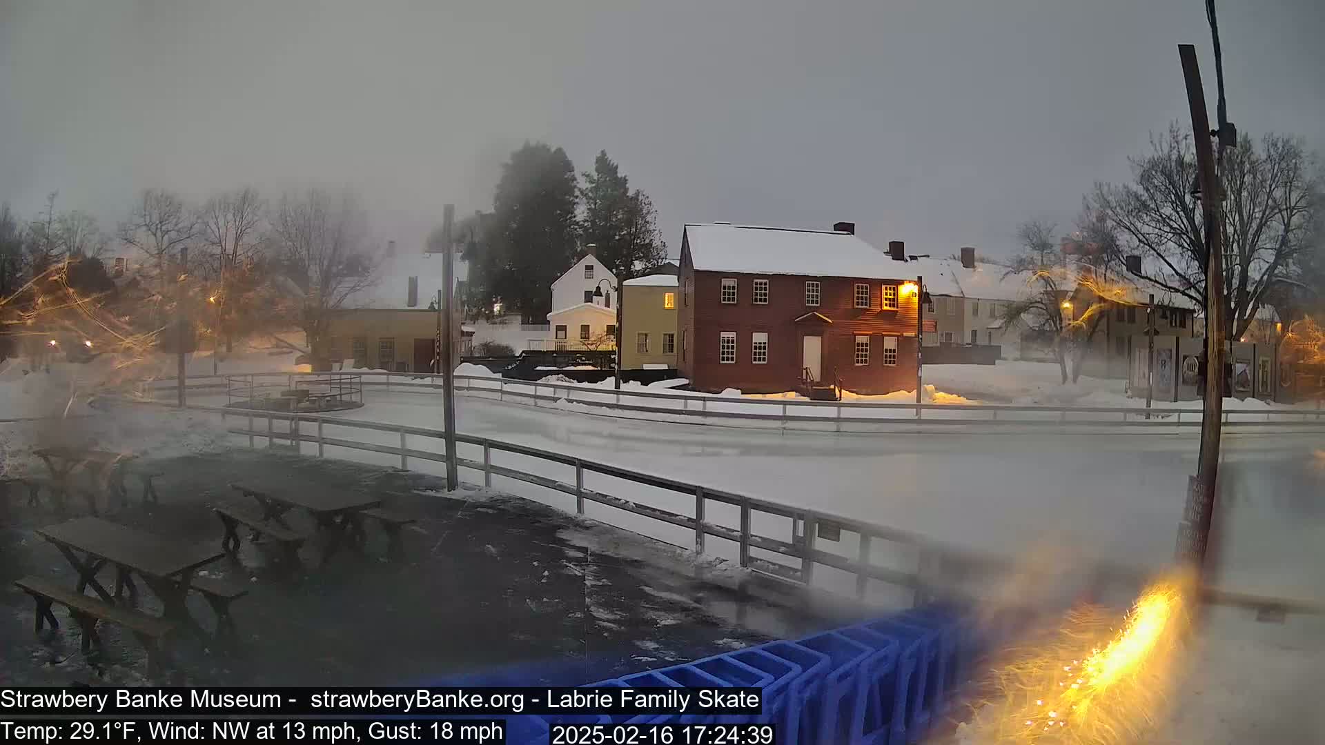A snow-covered outdoor ice rink is situated near several historic buildings under a cloudy, twilight sky with a northwest wind.