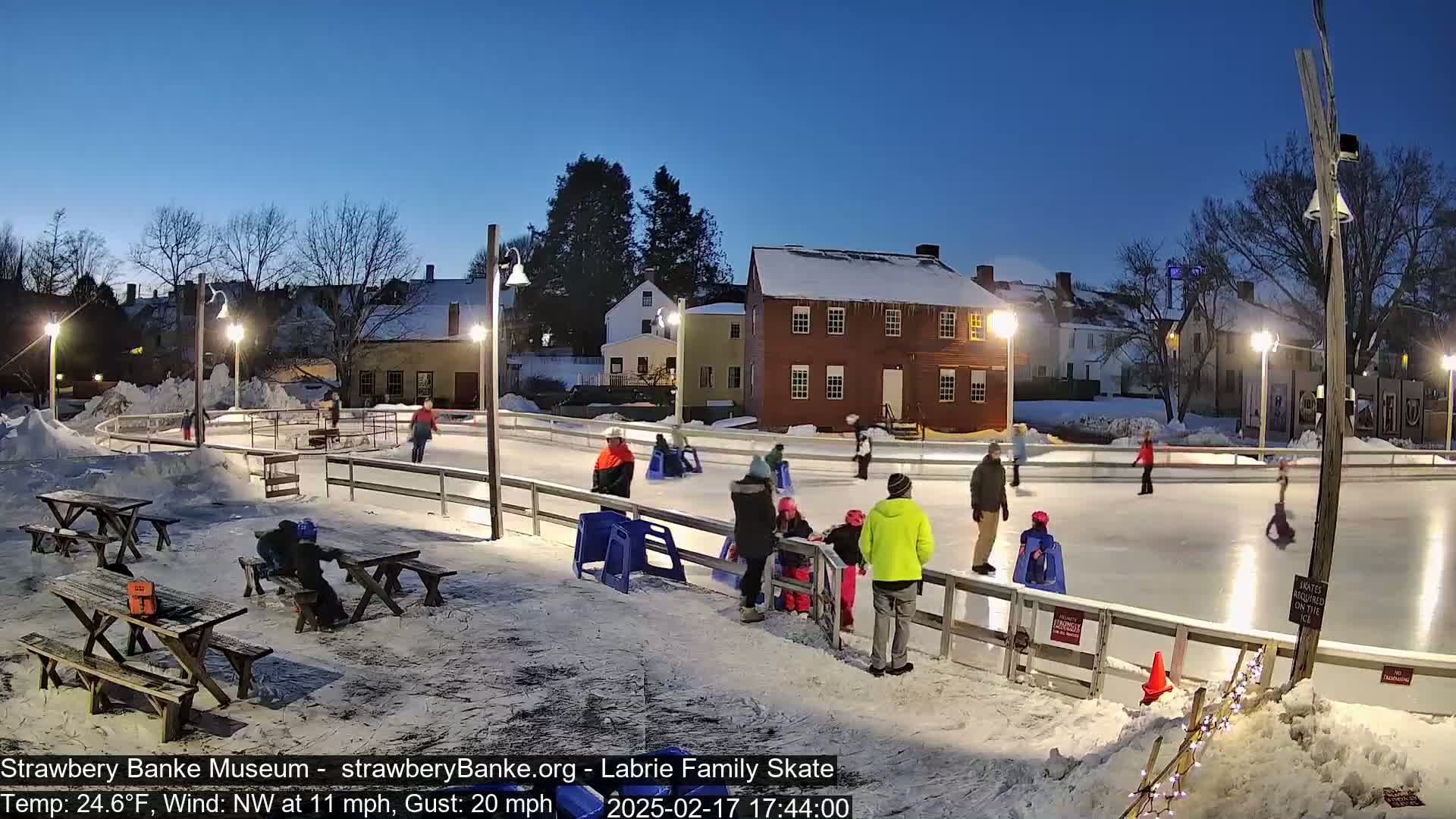 An outdoor ice skating rink, lit by lamps, is surrounded by snow-covered ground, picnic tables, and historic buildings under a twilight sky with a northwest wind.