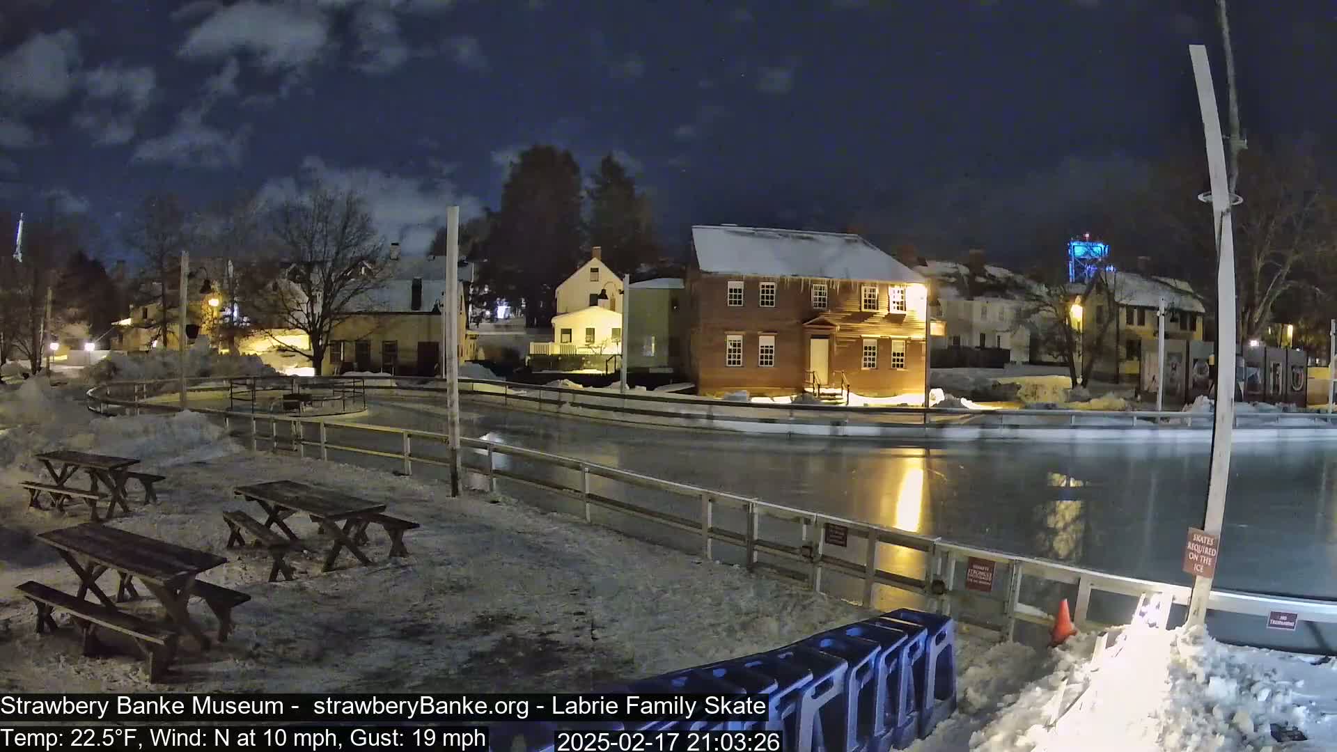 A nighttime view of an outdoor ice skating rink, surrounded by snow-covered picnic tables and historic buildings under a partly cloudy sky, with a moderate north wind.