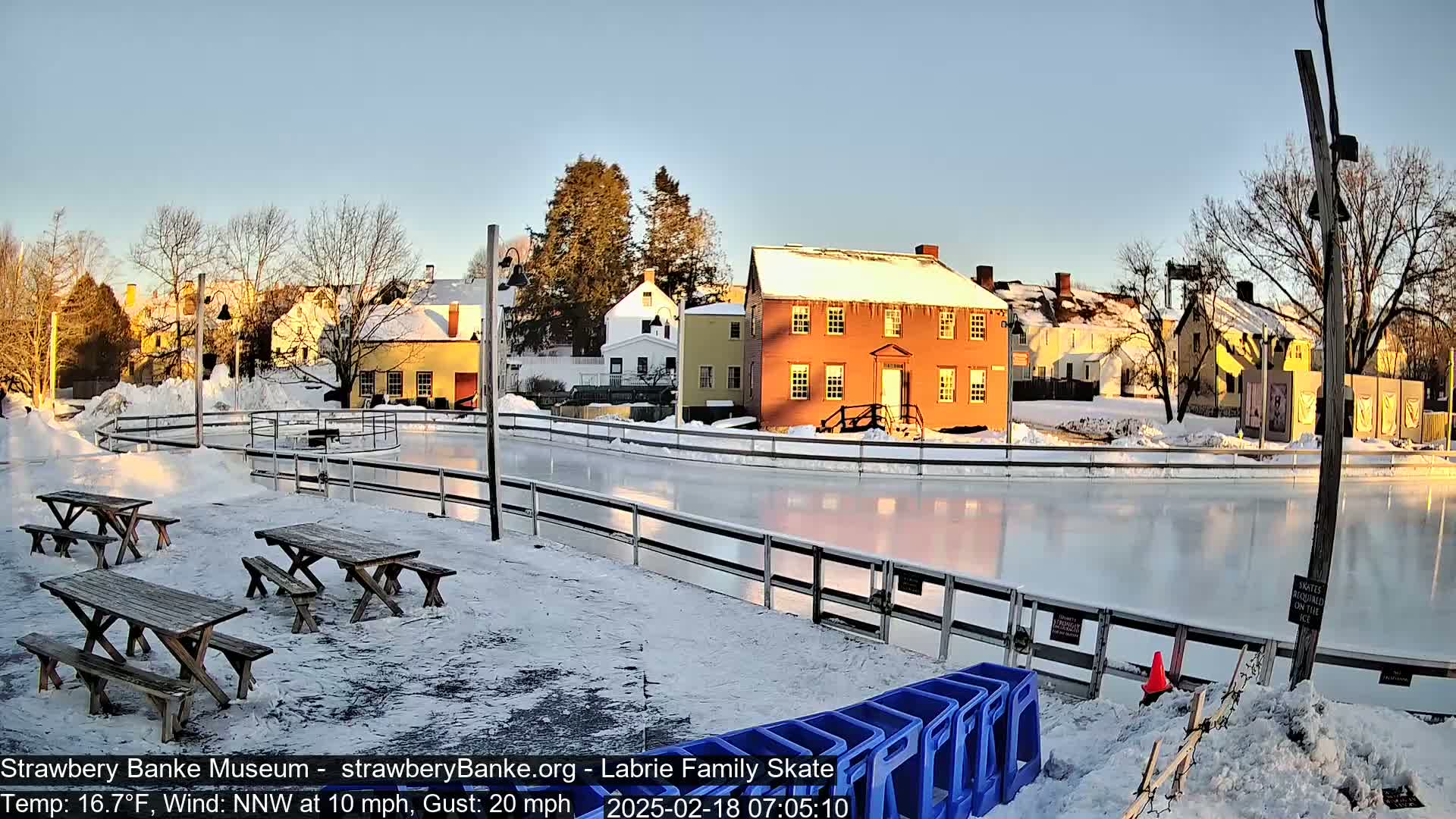 A frozen outdoor skating rink is surrounded by snow-covered picnic tables and historic buildings under a clear, sunny sky.