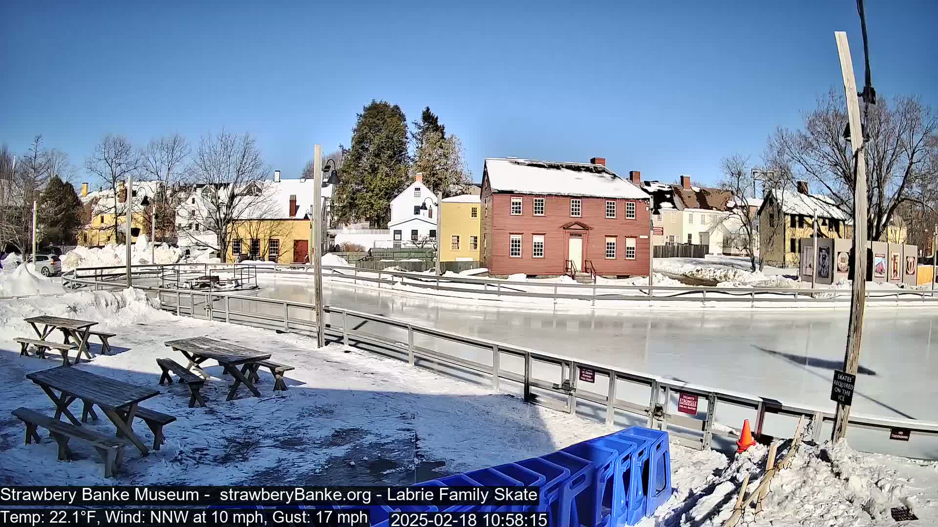 A snow-covered outdoor ice skating rink is situated next to several historic-looking buildings under a sunny, clear sky.