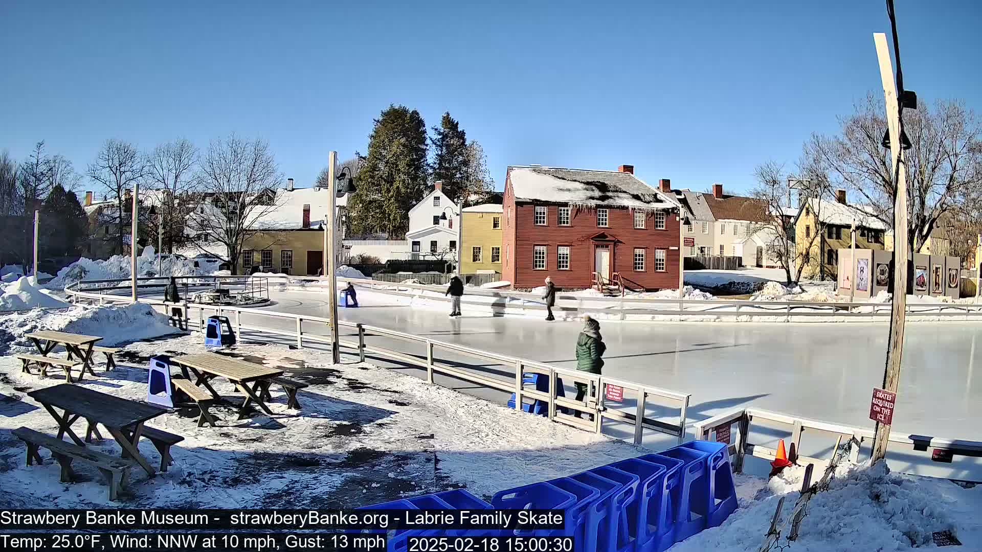 A sunny, cold day shows several people ice skating on an outdoor rink next to snow-covered picnic tables and historic buildings.