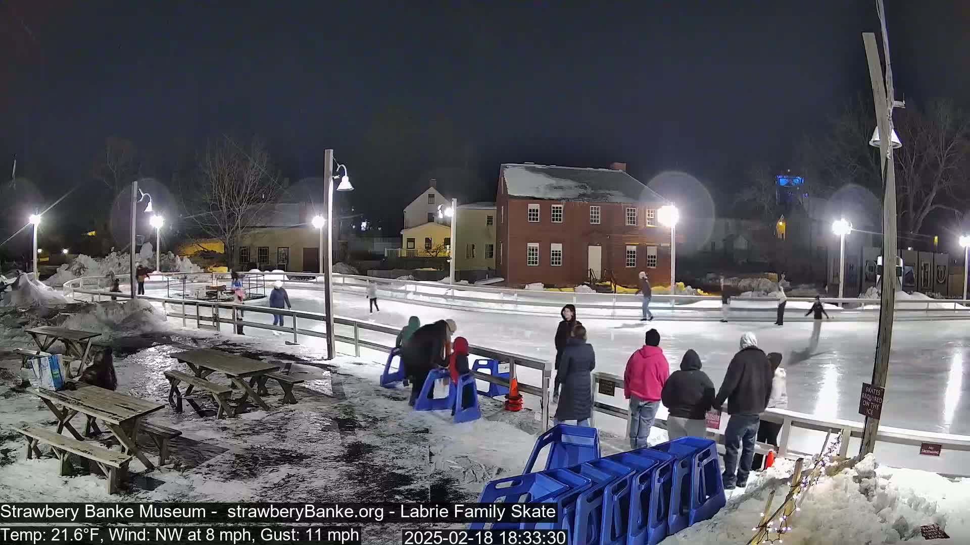 An outdoor ice skating rink at night, illuminated by lights, with several people ice skating and others standing nearby, in snowy conditions with a 21.6°F temperature and a northwest wind.