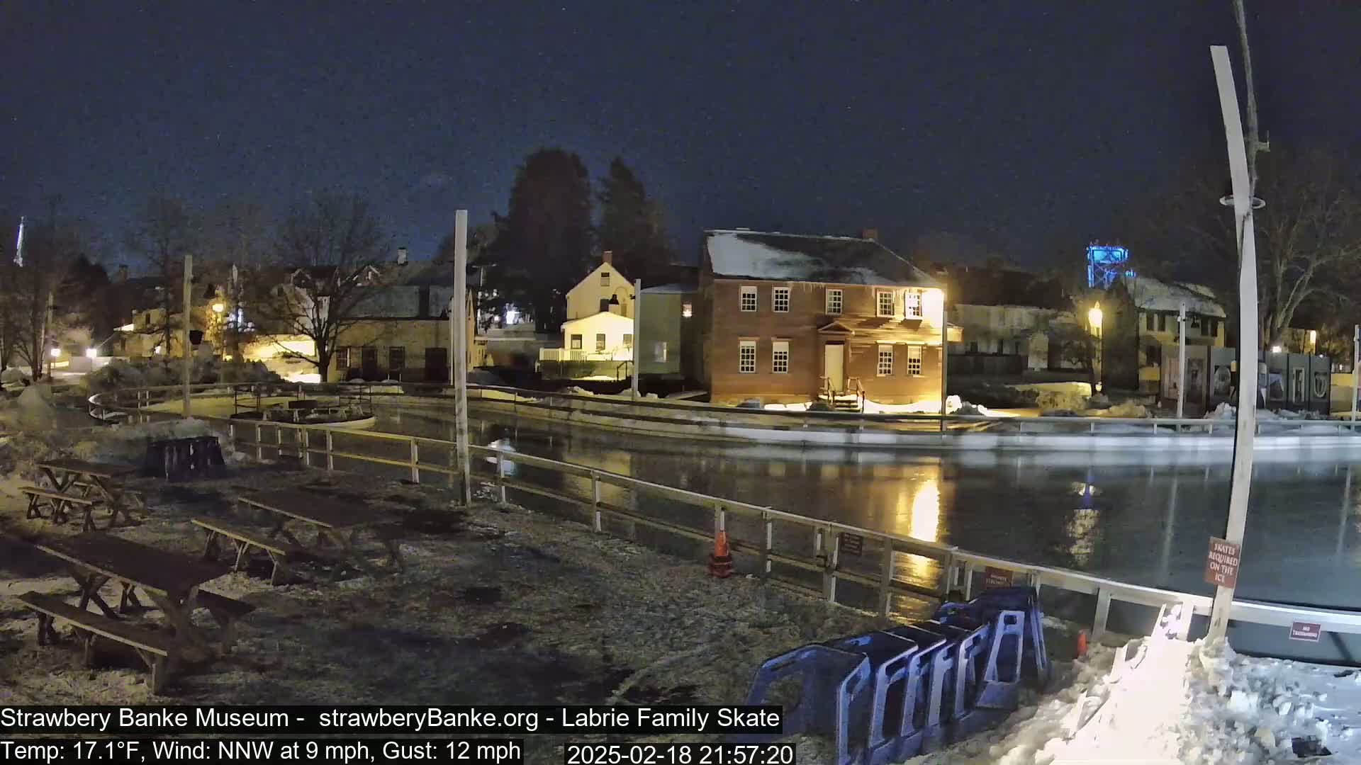 A nighttime view of a frozen outdoor skating rink next to several historic-looking buildings under a clear, starry sky.