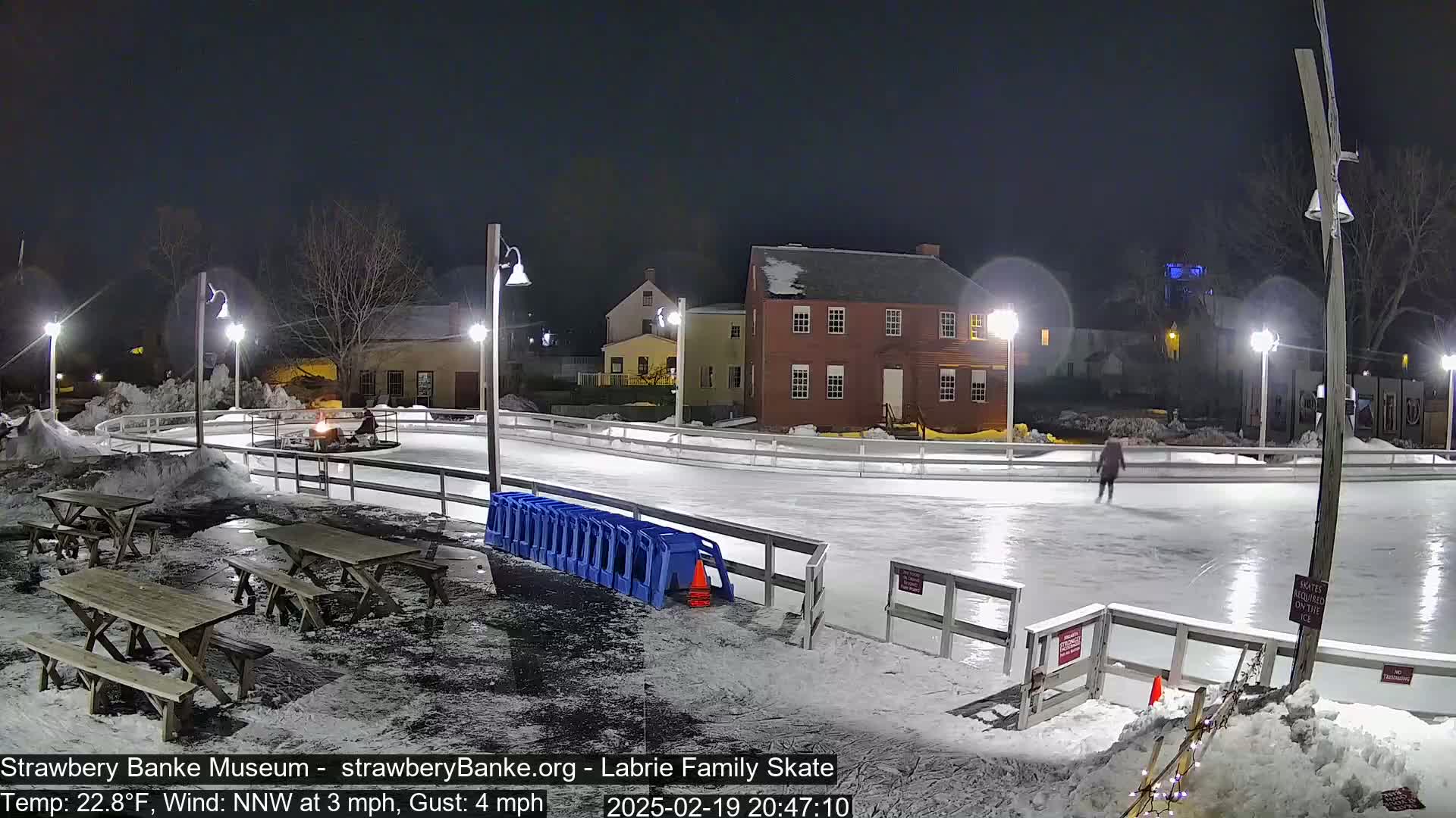 An outdoor ice skating rink at night, illuminated by lampposts, with a person skating, snow on the ground, and picnic tables nearby.