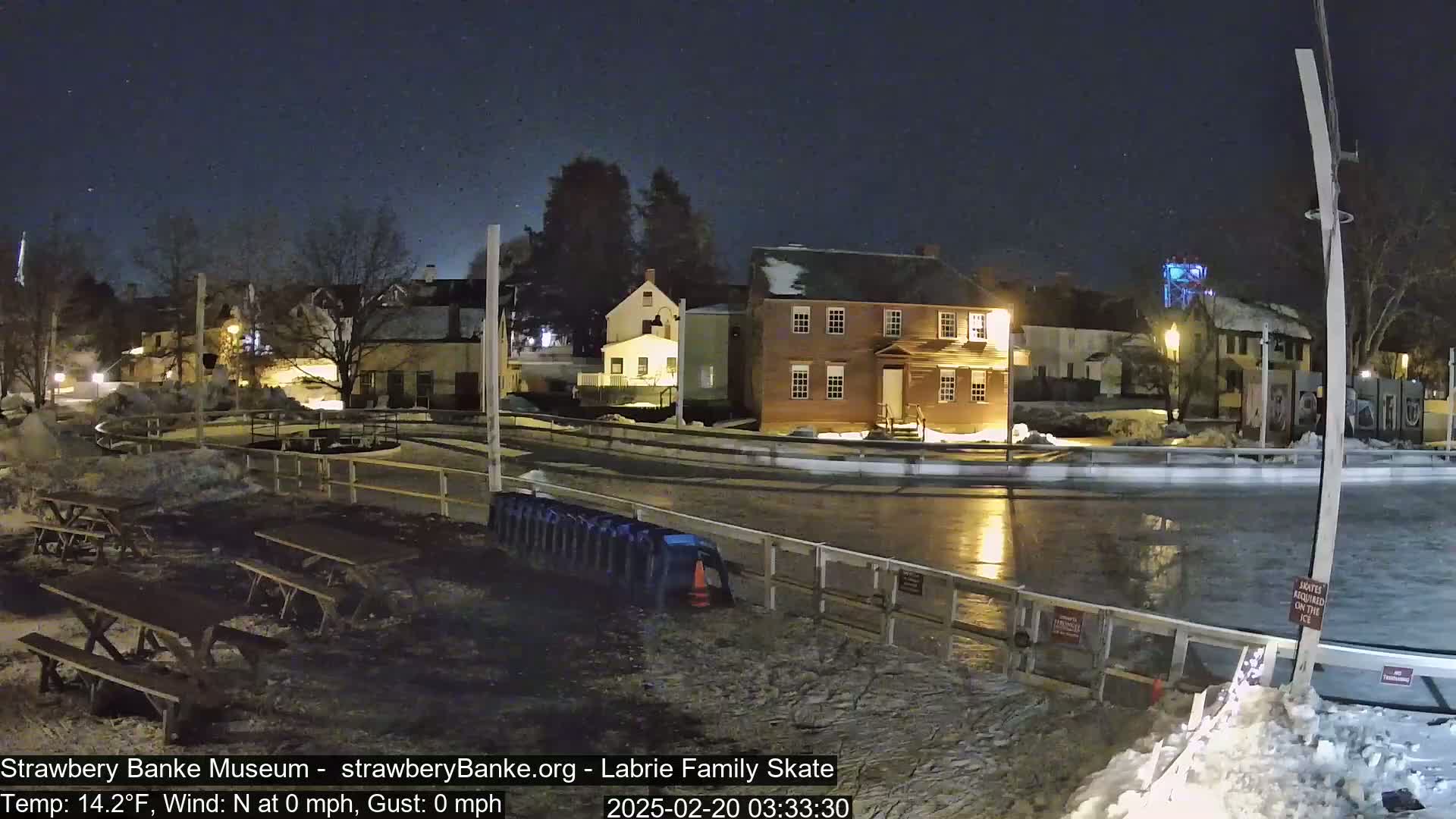 A nighttime view of a frozen outdoor skating rink next to several historic-looking buildings under a clear, dark sky.