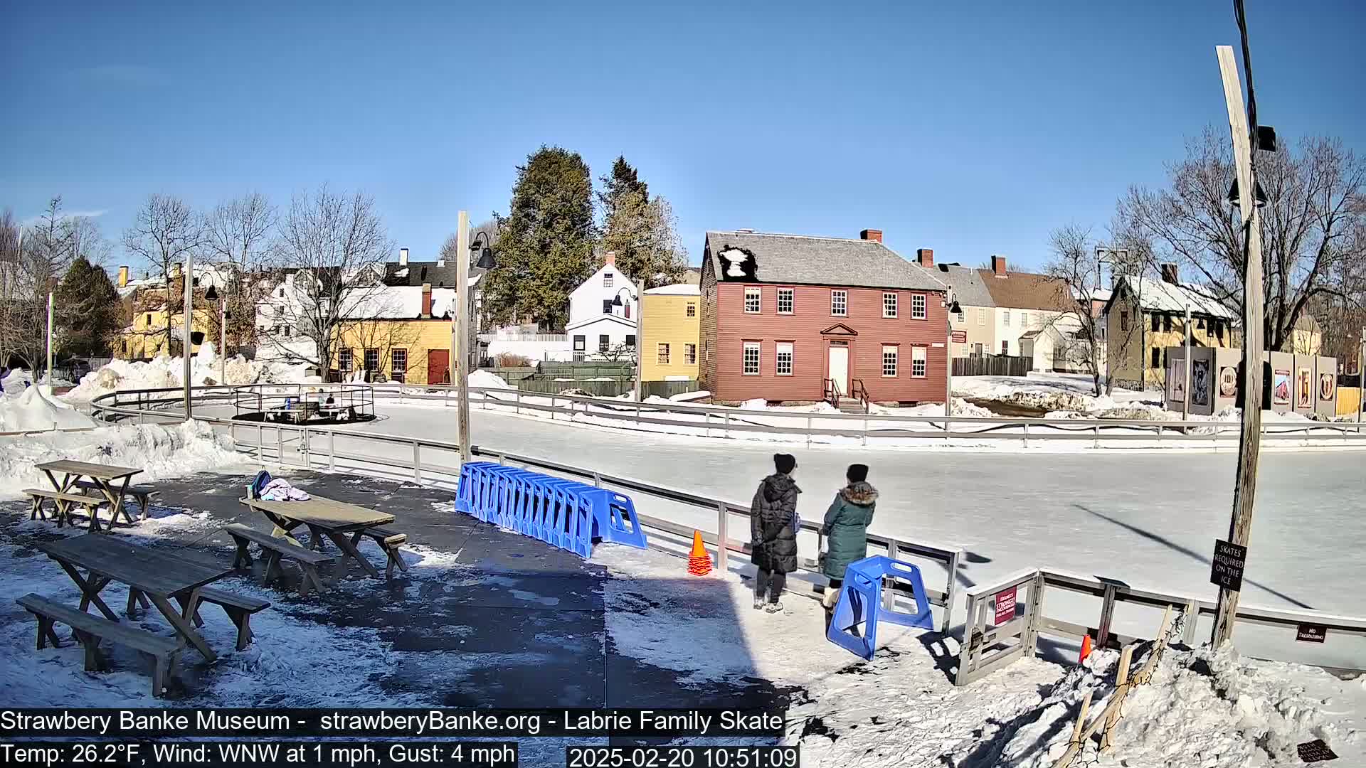 An outdoor ice skating rink is surrounded by snow-covered ground and historic-looking buildings under a sunny sky.