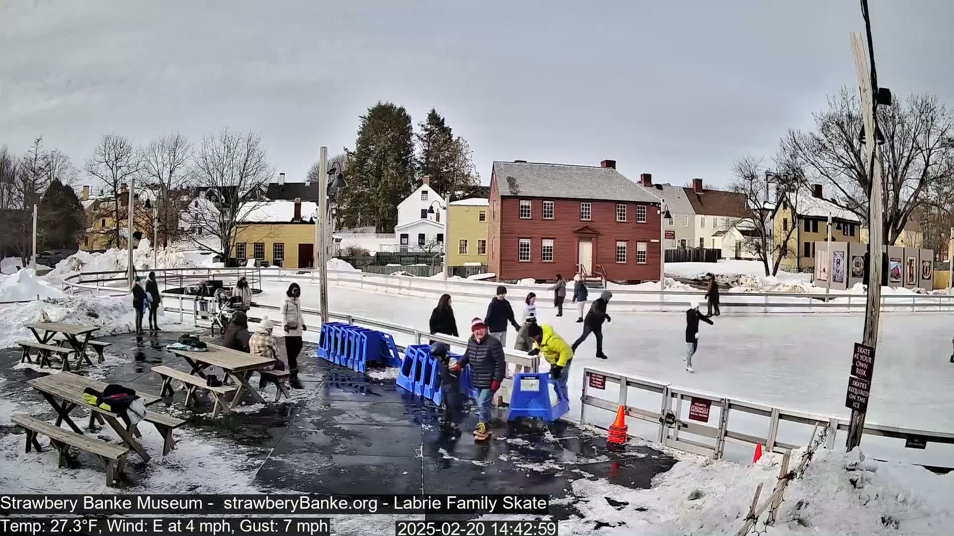 People ice skate on an outdoor rink surrounded by snow and historic buildings under a cloudy sky.
