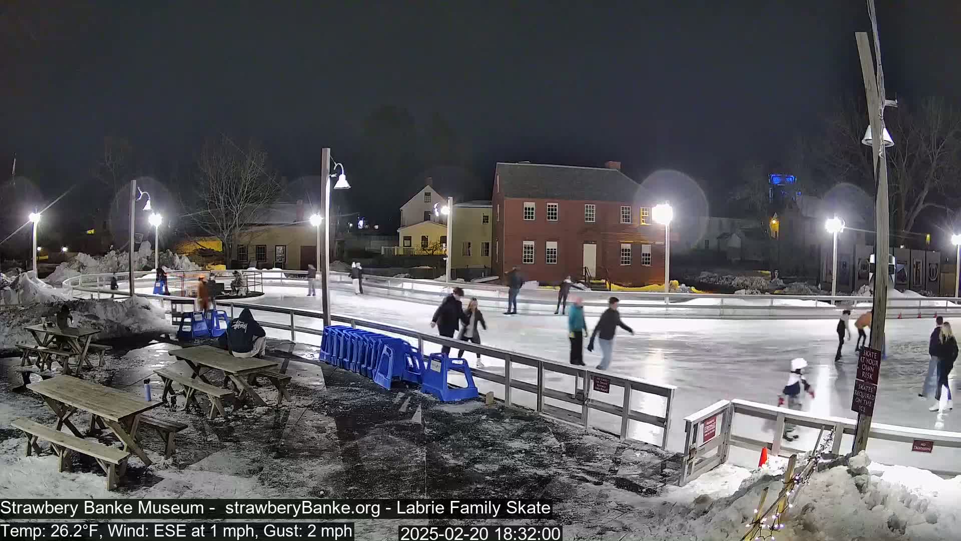 At night, several people ice skate on an outdoor rink illuminated by lampposts, with snow on the ground and nearby picnic tables.