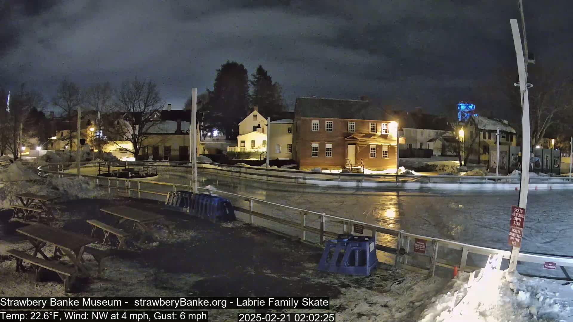 A nighttime view of an outdoor ice skating rink surrounded by snow and historic buildings under a cloudy sky with a northwest wind.