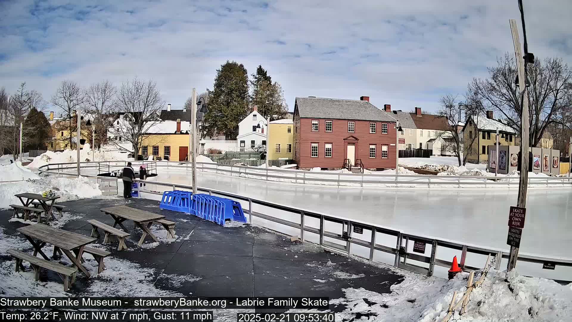 An outdoor ice skating rink, surrounded by snow and historic-looking buildings, under a partly sunny sky.