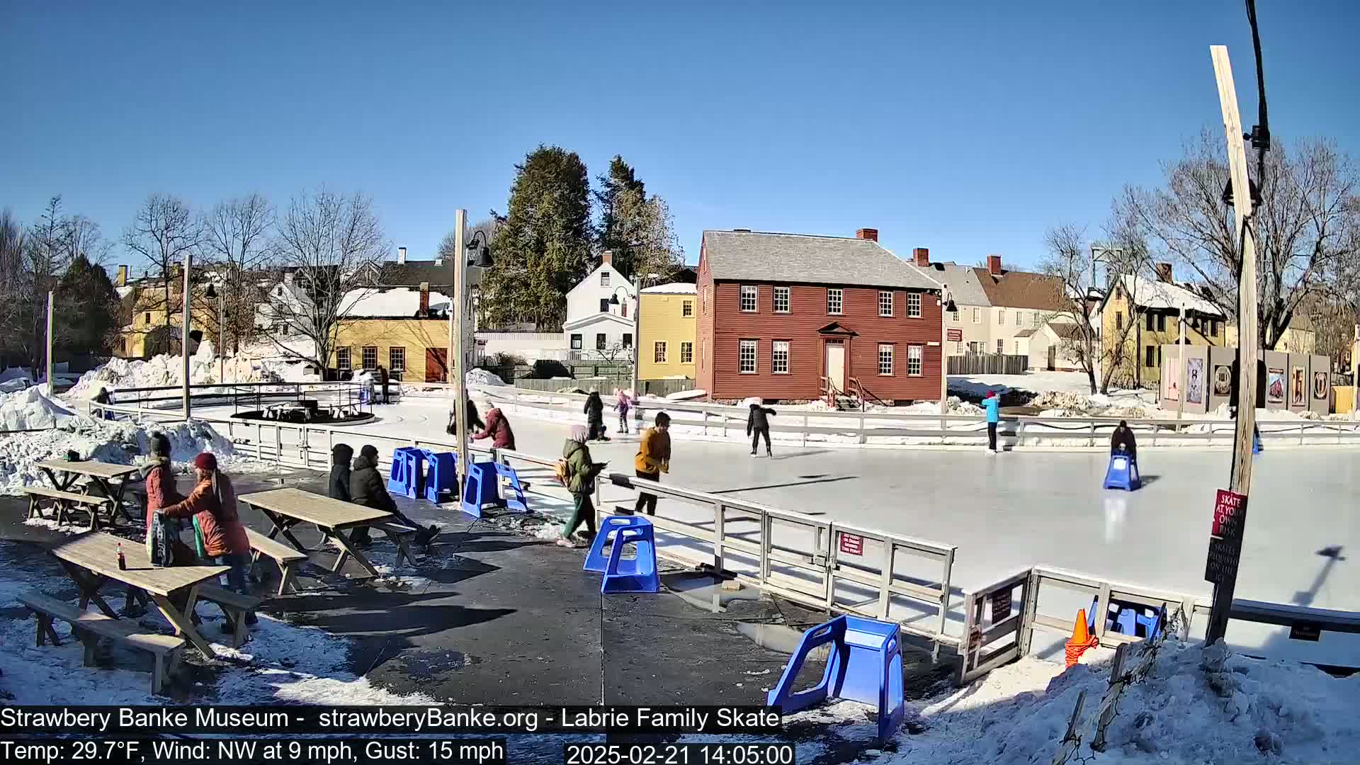 An outdoor ice skating rink with several people skating and others sitting at picnic tables, surrounded by historic-looking buildings under a sunny sky.