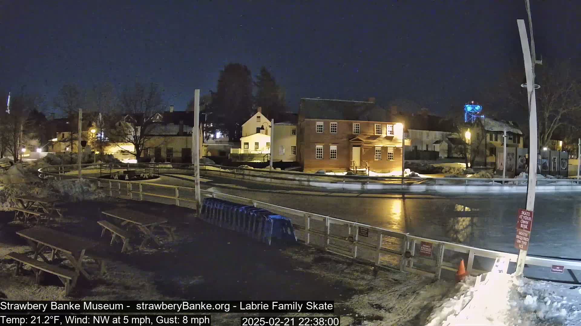 A nighttime view of an outdoor ice skating rink with surrounding buildings under a clear, starry sky, and a light dusting of snow on the ground.