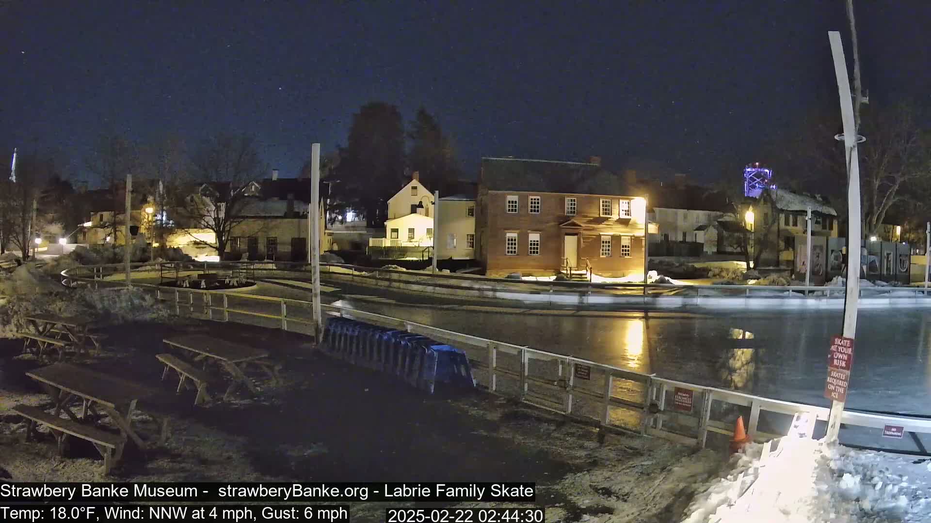A nighttime view of an outdoor ice skating rink, surrounded by snow and historic-looking buildings, under a clear, starry sky with a light northwest wind.