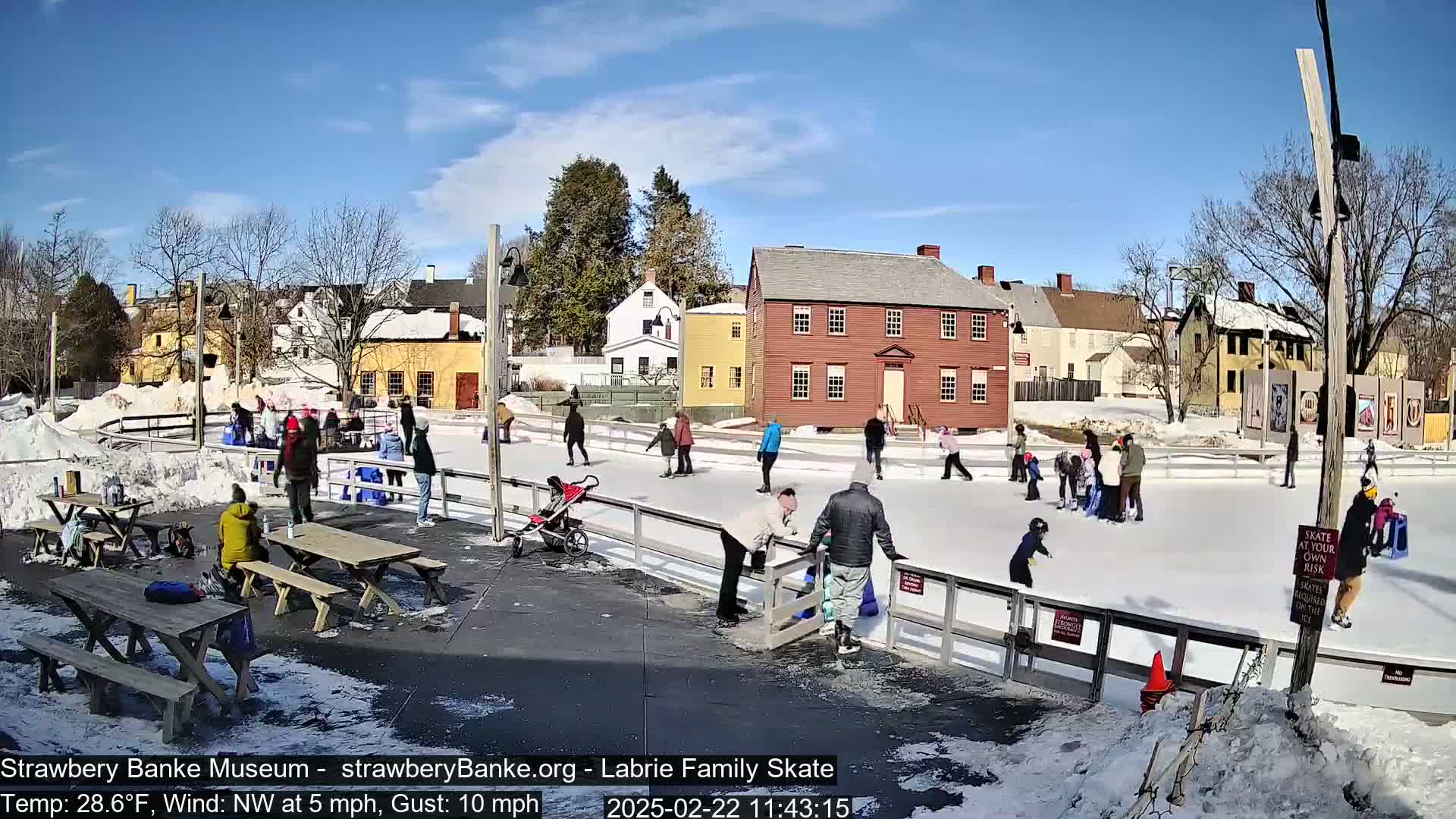 A sunny day shows people ice skating on an outdoor rink, surrounded by historic buildings and picnic tables.