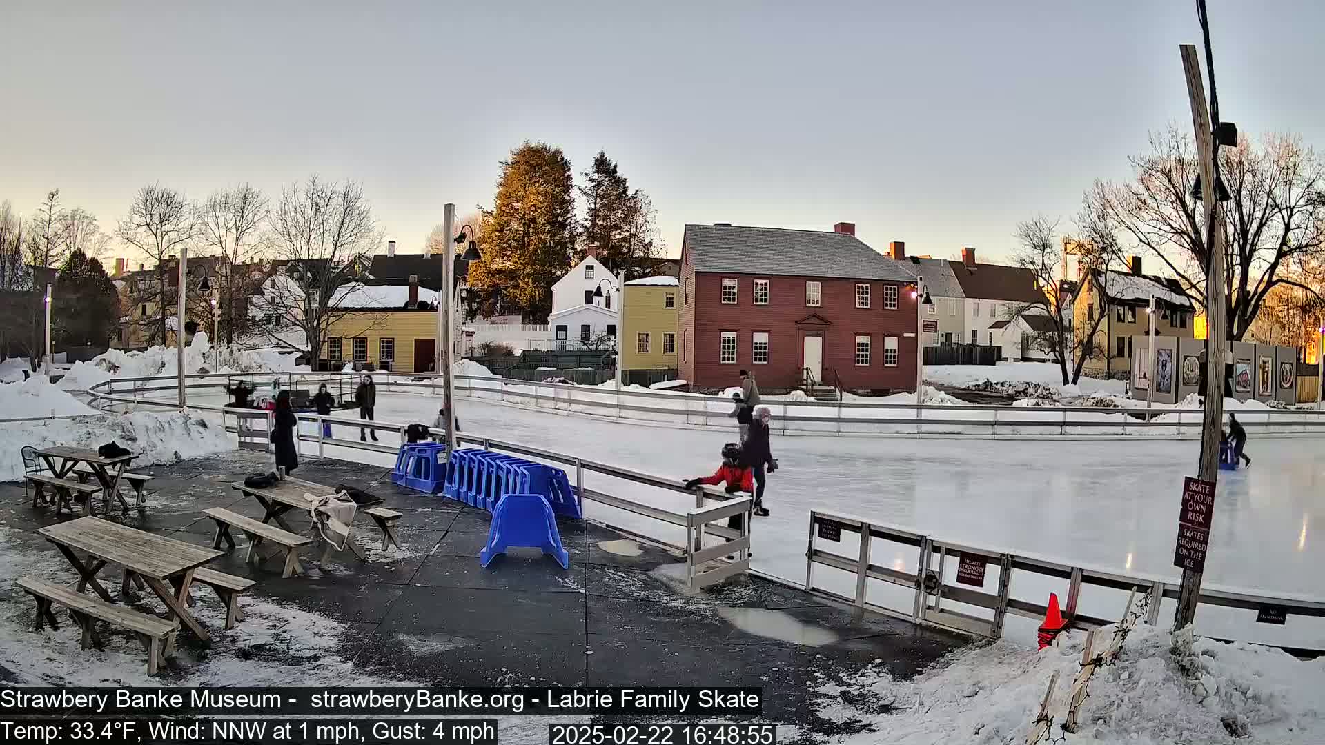 An outdoor ice skating rink, surrounded by snow and historic buildings, with several people ice skating and others seated at picnic tables nearby under a twilight sky.
