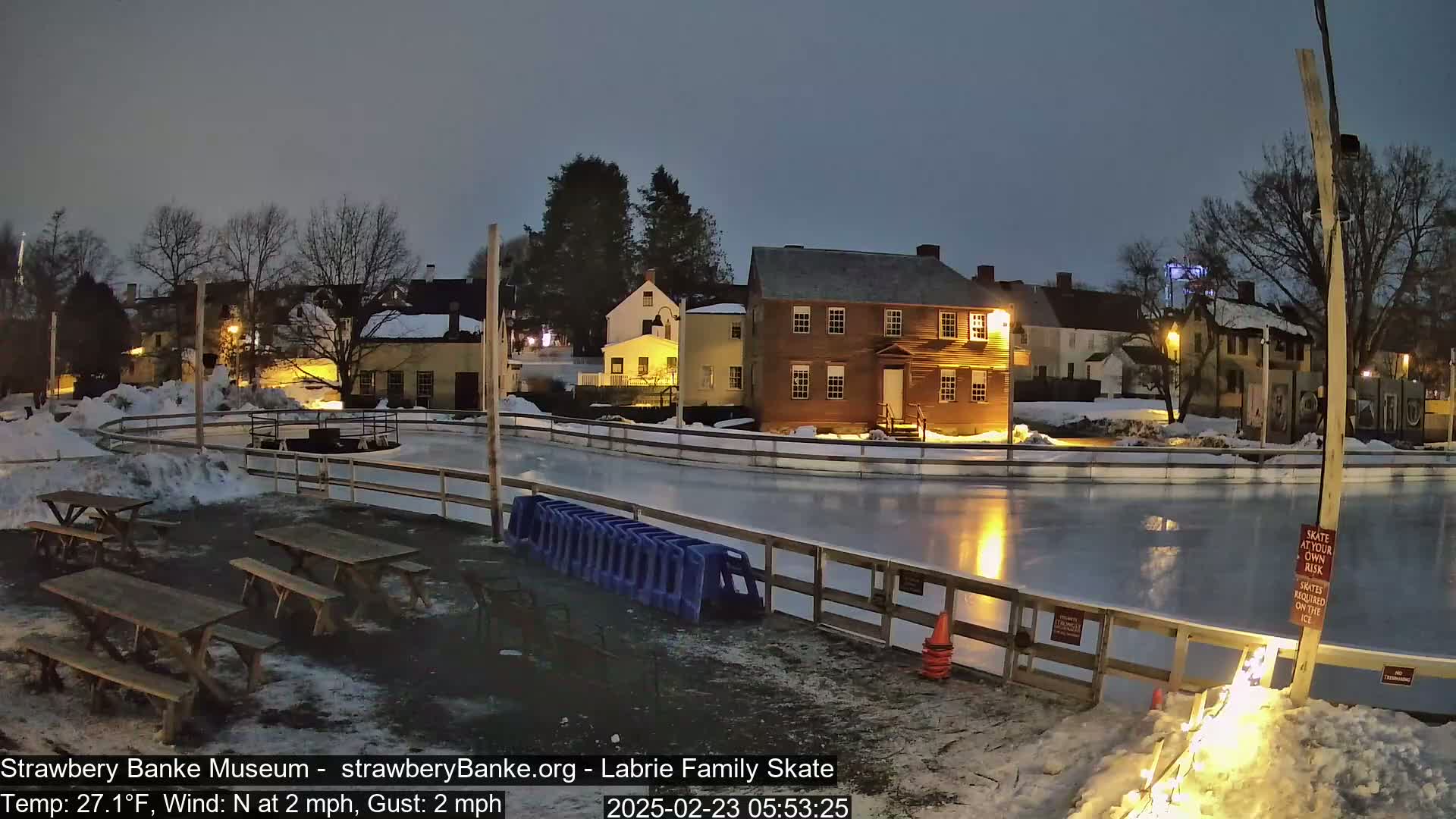 A nighttime view of an outdoor ice skating rink, surrounded by snow and historic-looking buildings, under a dark, clear sky.