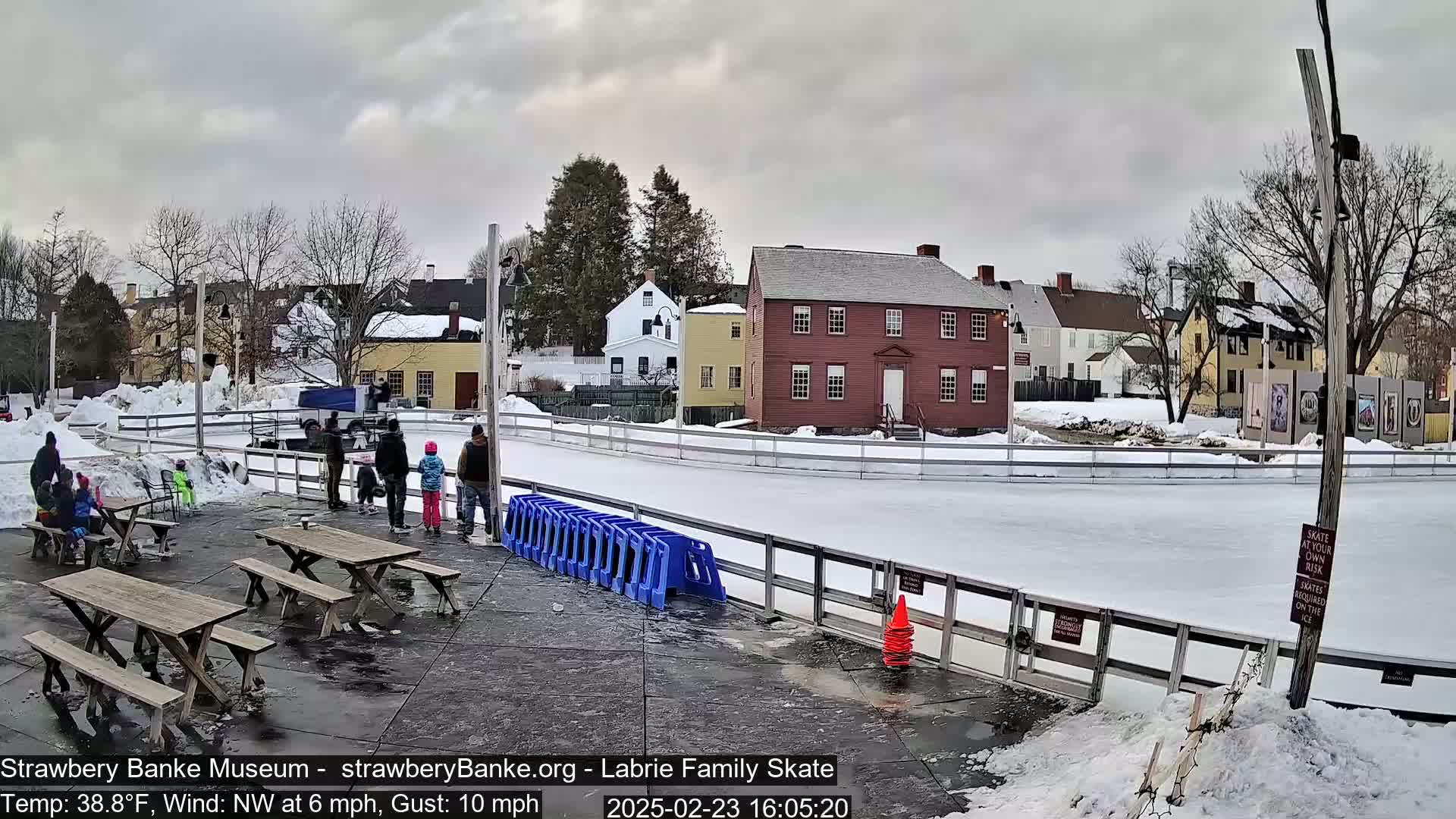 An outdoor ice skating rink with people skating and sitting at picnic tables, surrounded by snow-covered ground and historical-looking buildings under an overcast sky.