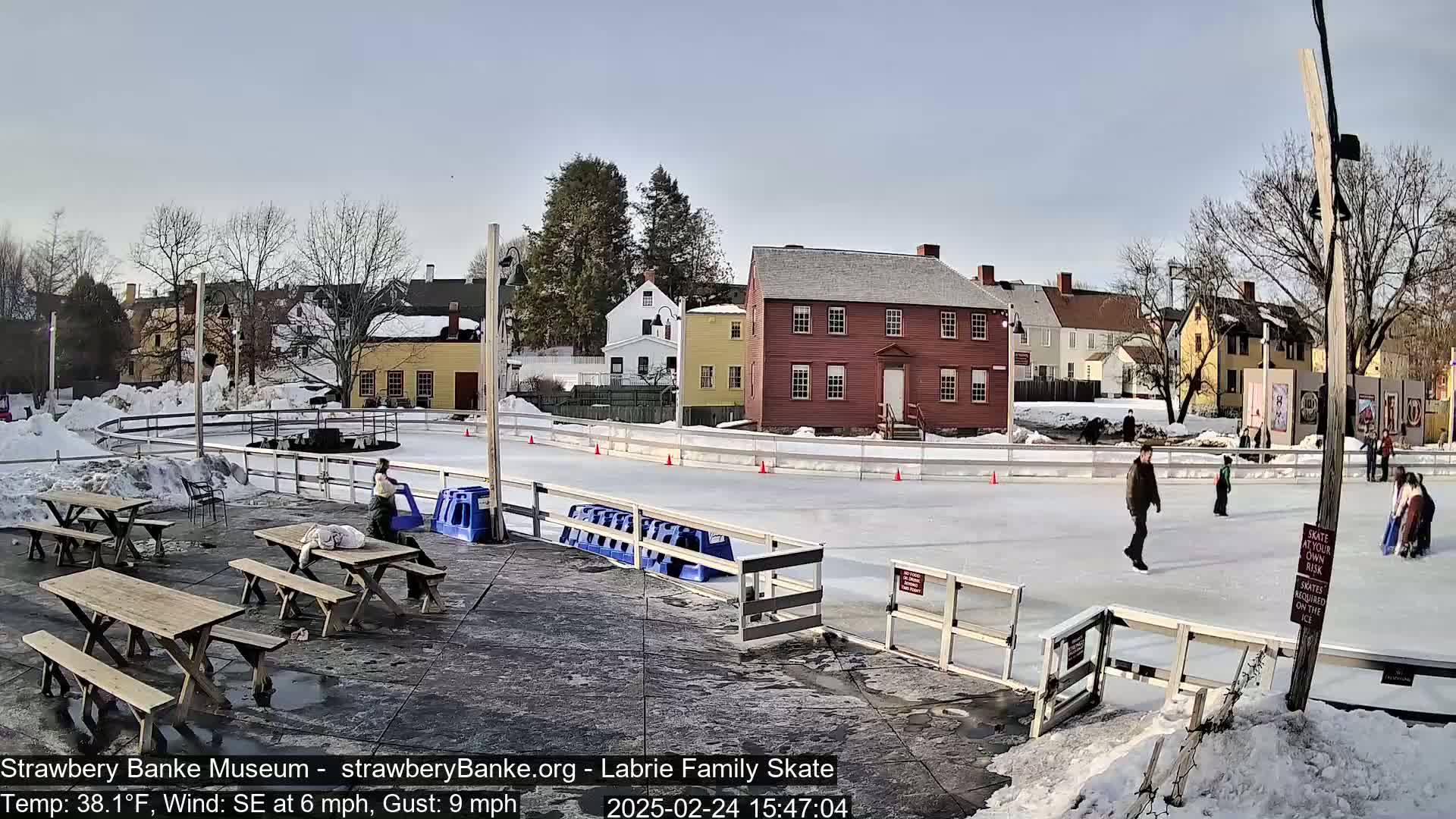 An outdoor ice-skating rink surrounded by snow and historic buildings, with several people ice skating and picnic tables nearby under a partly sunny sky.