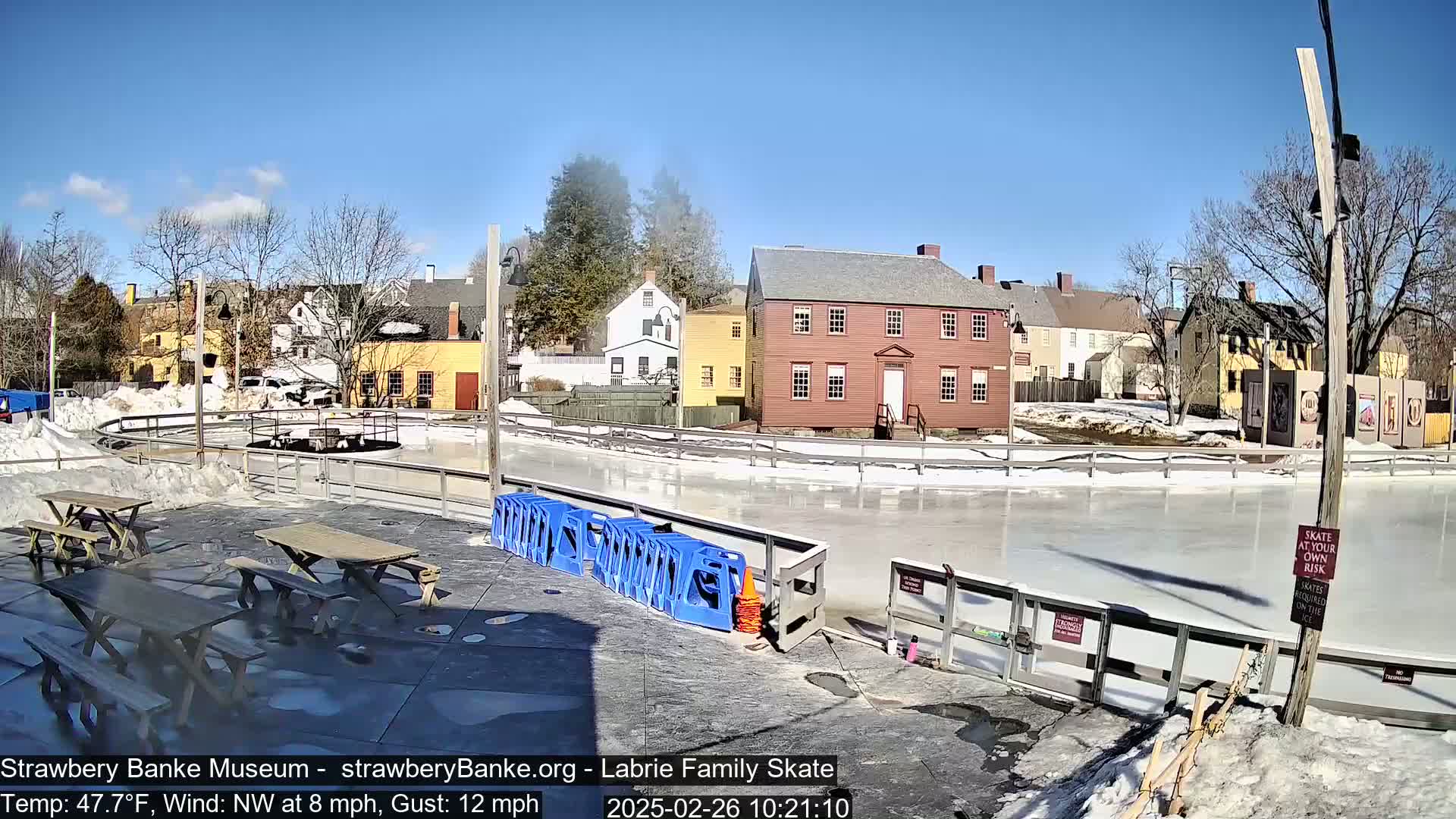An outdoor ice rink next to several historic-looking buildings on a sunny day with a northwest wind.