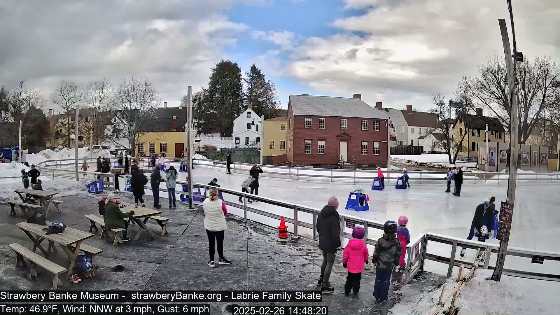 People ice skate on an outdoor rink next to picnic tables and historic buildings under a partly cloudy sky.