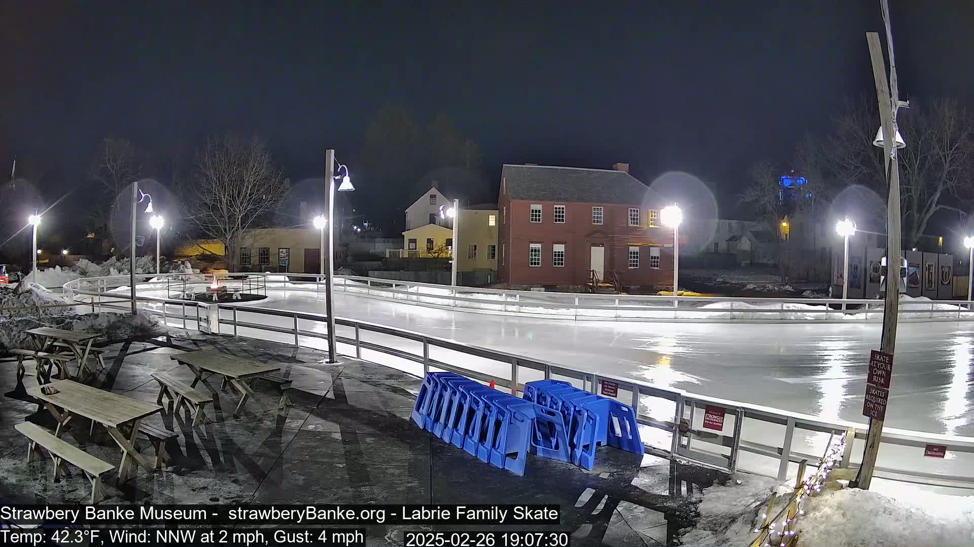 An outdoor ice rink at night, lit by several lampposts, is surrounded by buildings and a few picnic tables, with a light NNW wind.