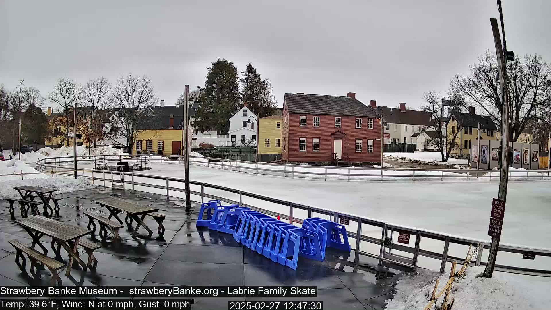 An outdoor ice skating rink is situated near several historic buildings on a partly snowy day under an overcast sky.