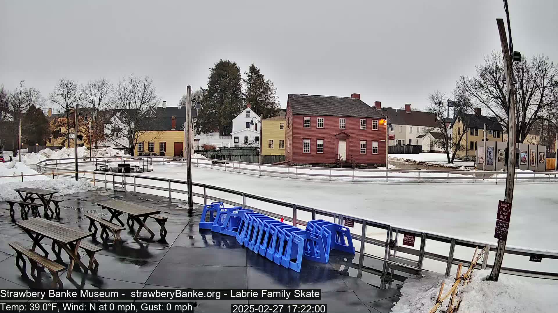 An outdoor ice skating rink, surrounded by historical buildings and picnic tables, on an overcast day.