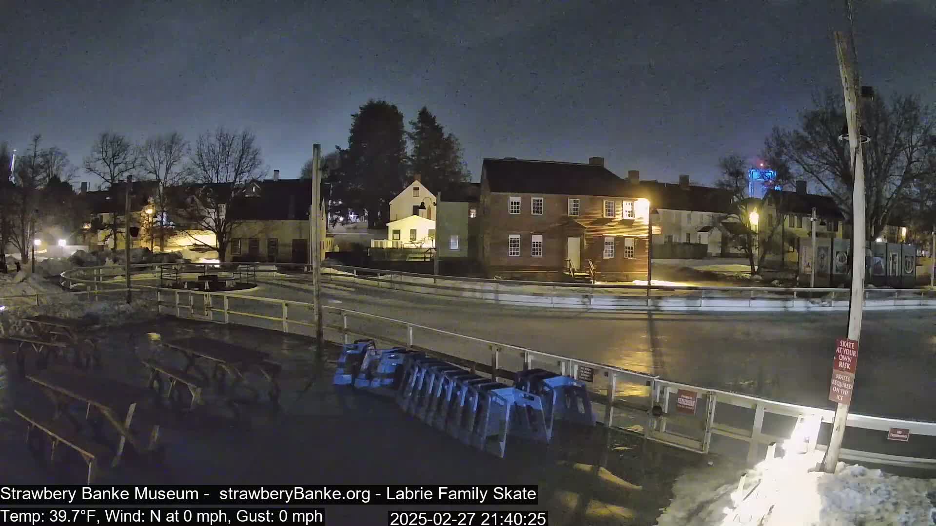 A nighttime view of a mostly empty outdoor ice rink near several historic-looking buildings under a dark, clear sky.