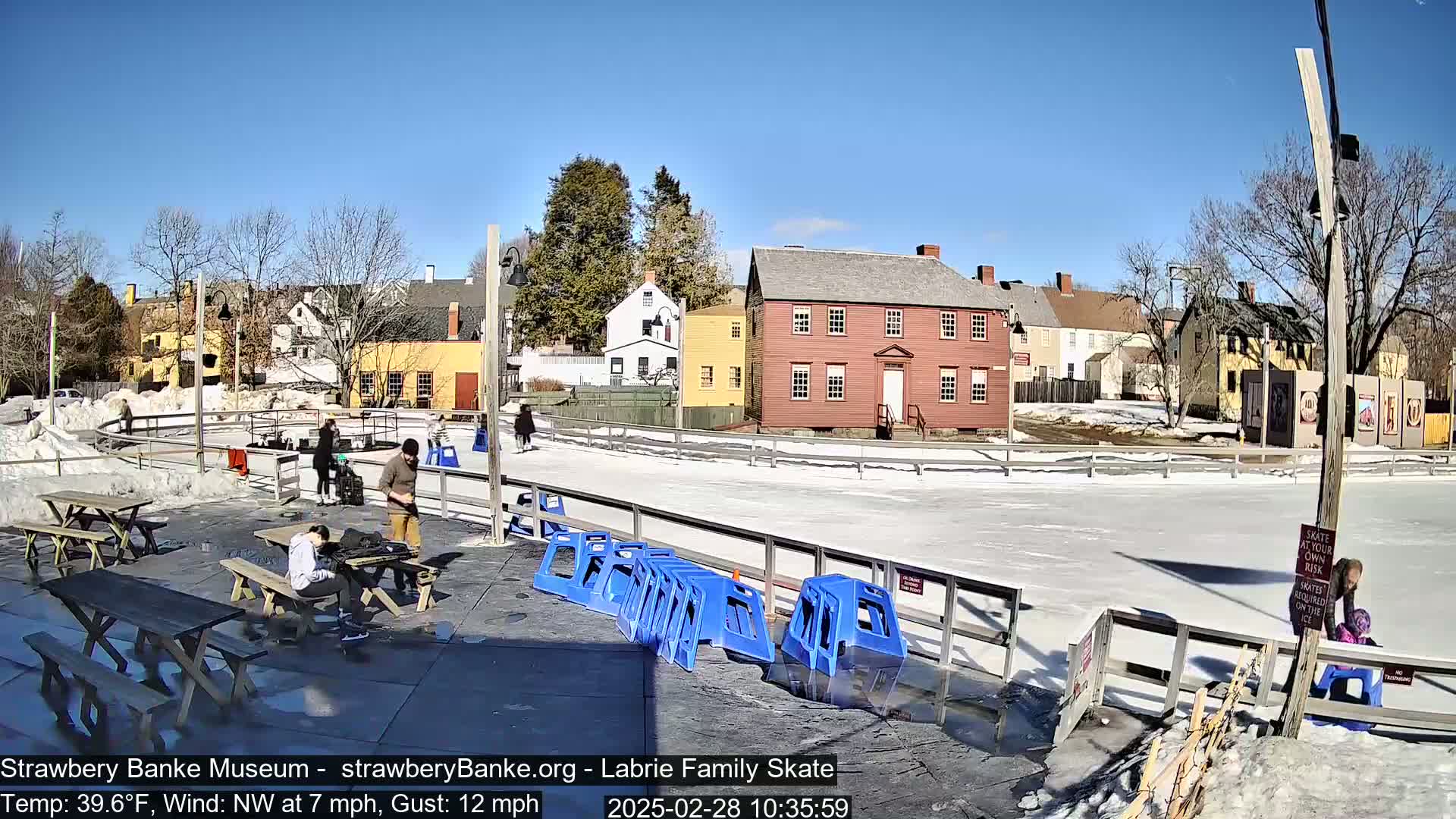 A sunny outdoor ice skating rink with a few people skating and others sitting at picnic tables, surrounded by historic-looking buildings.