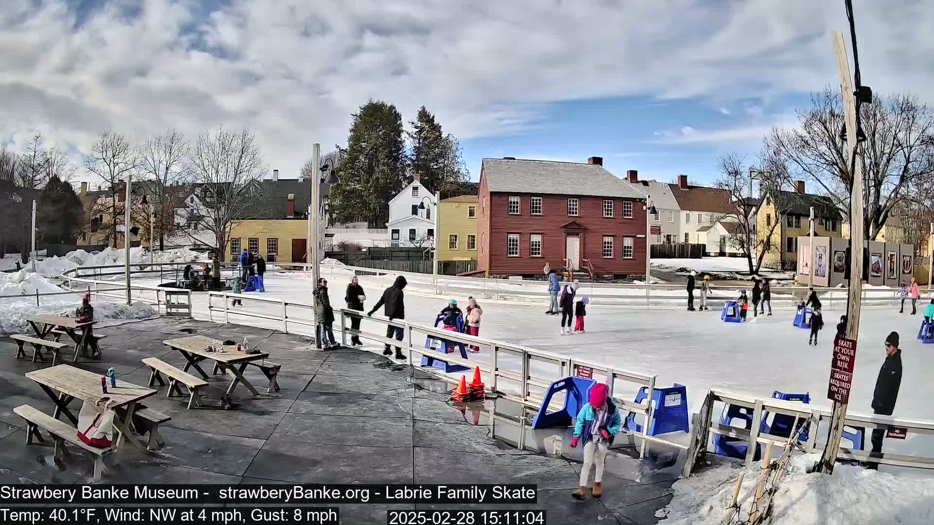 An outdoor ice skating rink, surrounded by historic buildings, is populated with skaters and onlookers on a partly sunny day with a northwest wind.