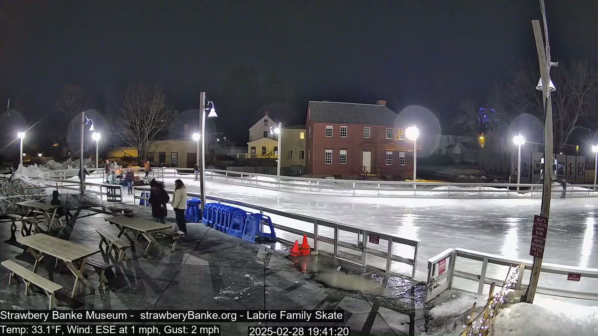 An outdoor ice skating rink at night, lit by several lamps, with a few people skating and others standing nearby, under a clear, dark sky and a 33°F temperature with an ESE wind.