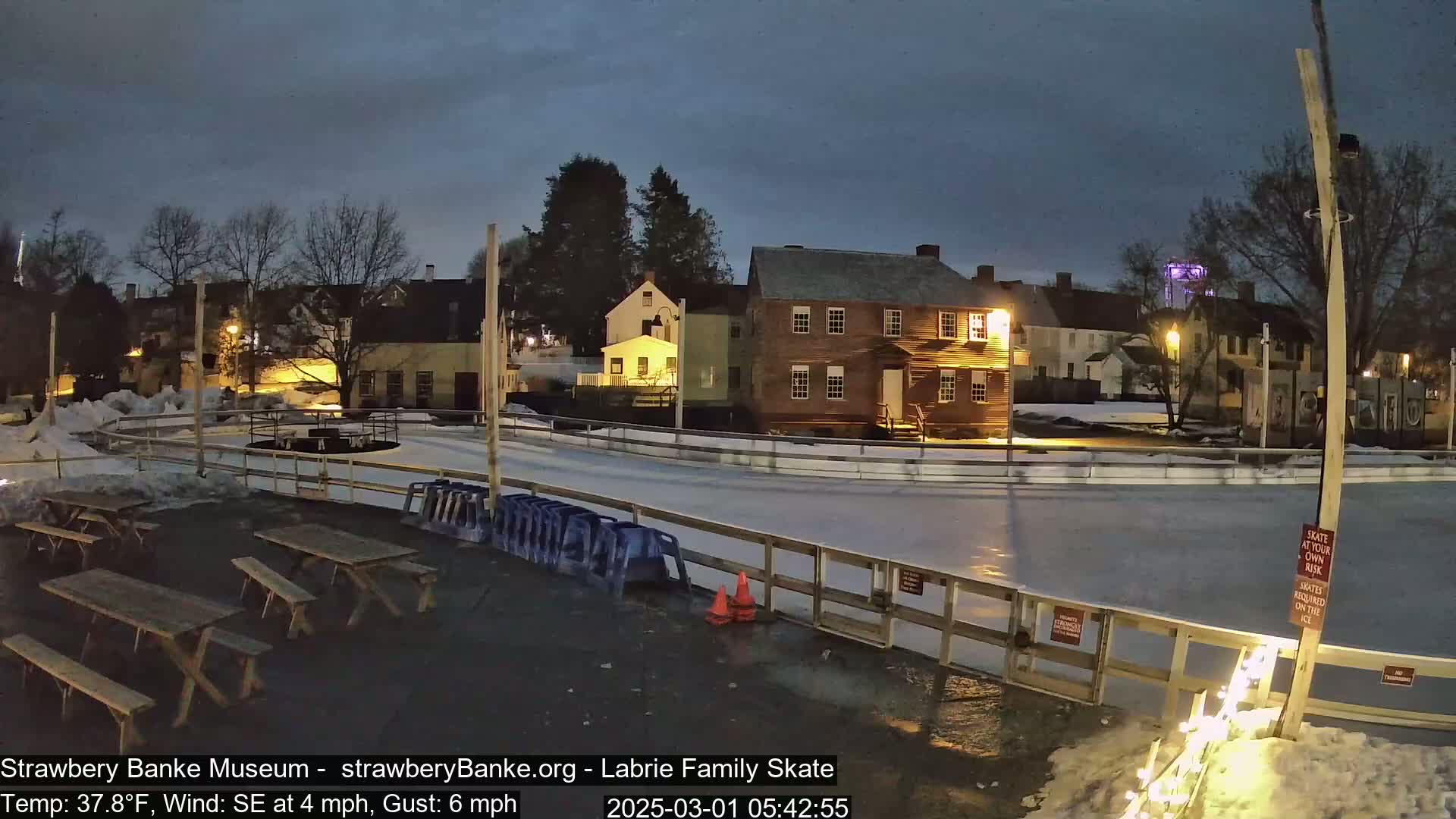 An outdoor ice skating rink, partially lit, is situated near several historic-looking buildings under a dark, twilight sky with a southeast wind.