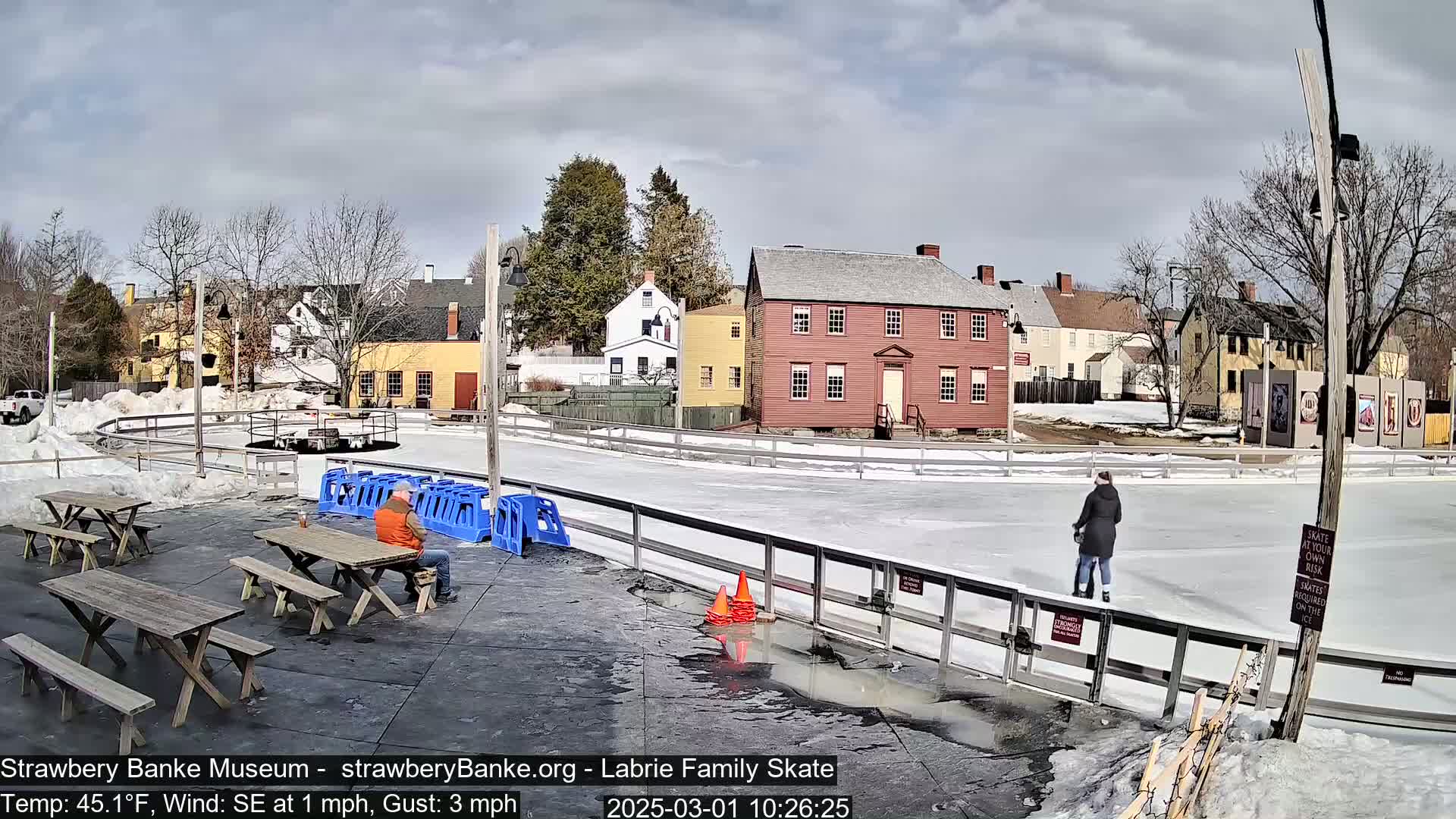 An outdoor ice skating rink, partially melted in patches, sits beside several wooden picnic tables with a person sitting at one and another person ice skating, all in front of a row of historic-looking buildings under a partly cloudy sky.