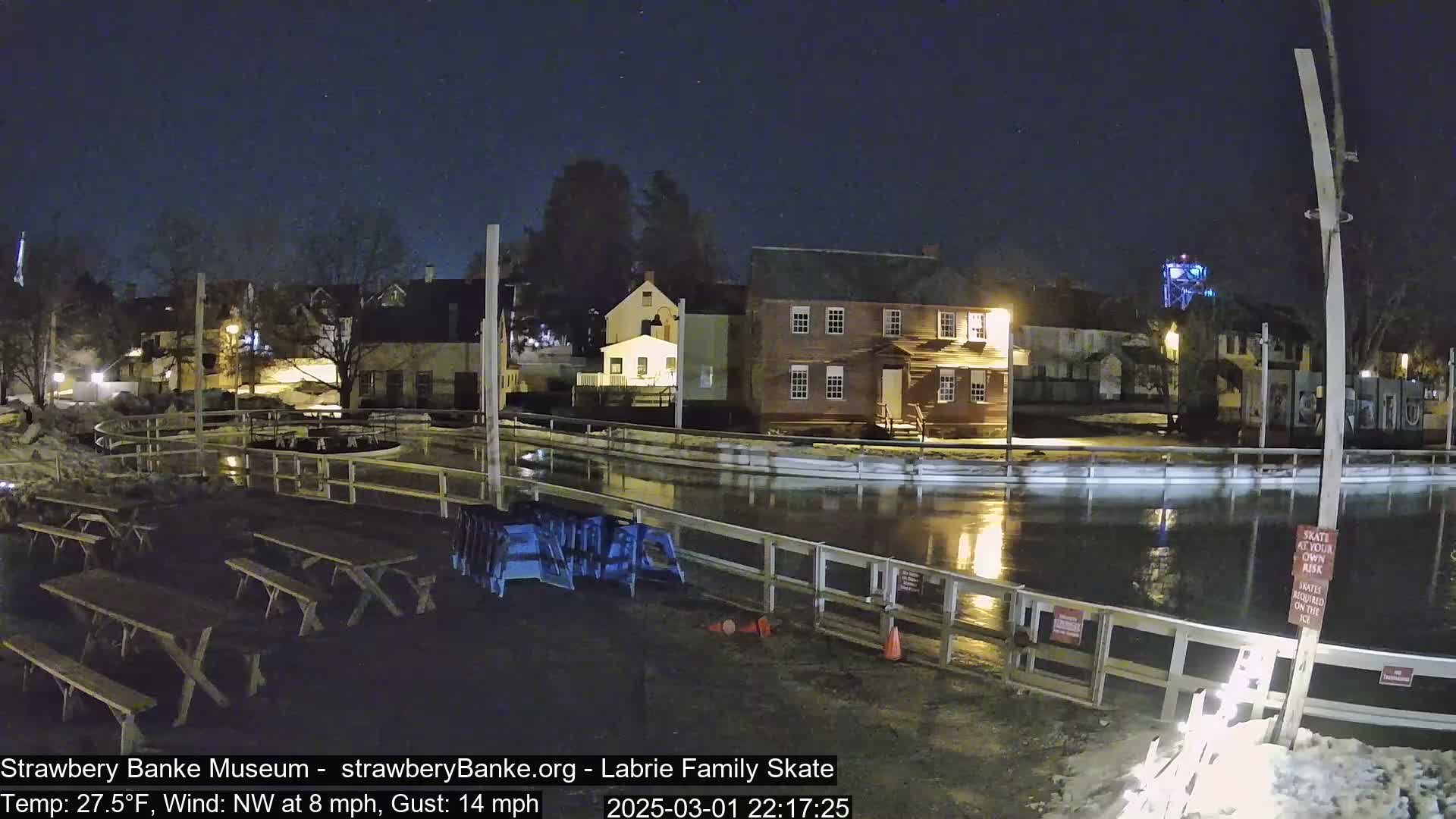A nighttime view of an outdoor ice rink surrounded by buildings, with picnic tables nearby under a clear, dark sky.