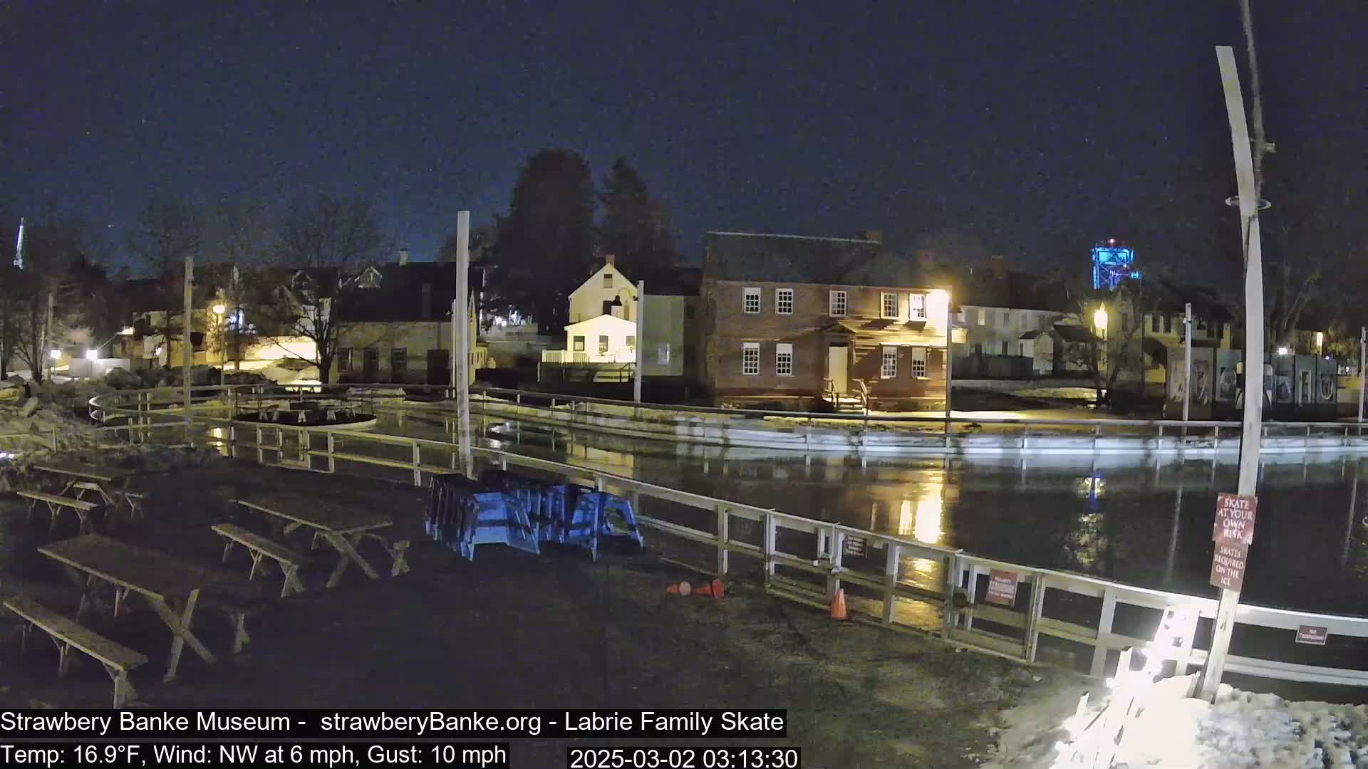 A nighttime view of a flooded outdoor ice rink next to several buildings under a clear, dark sky.