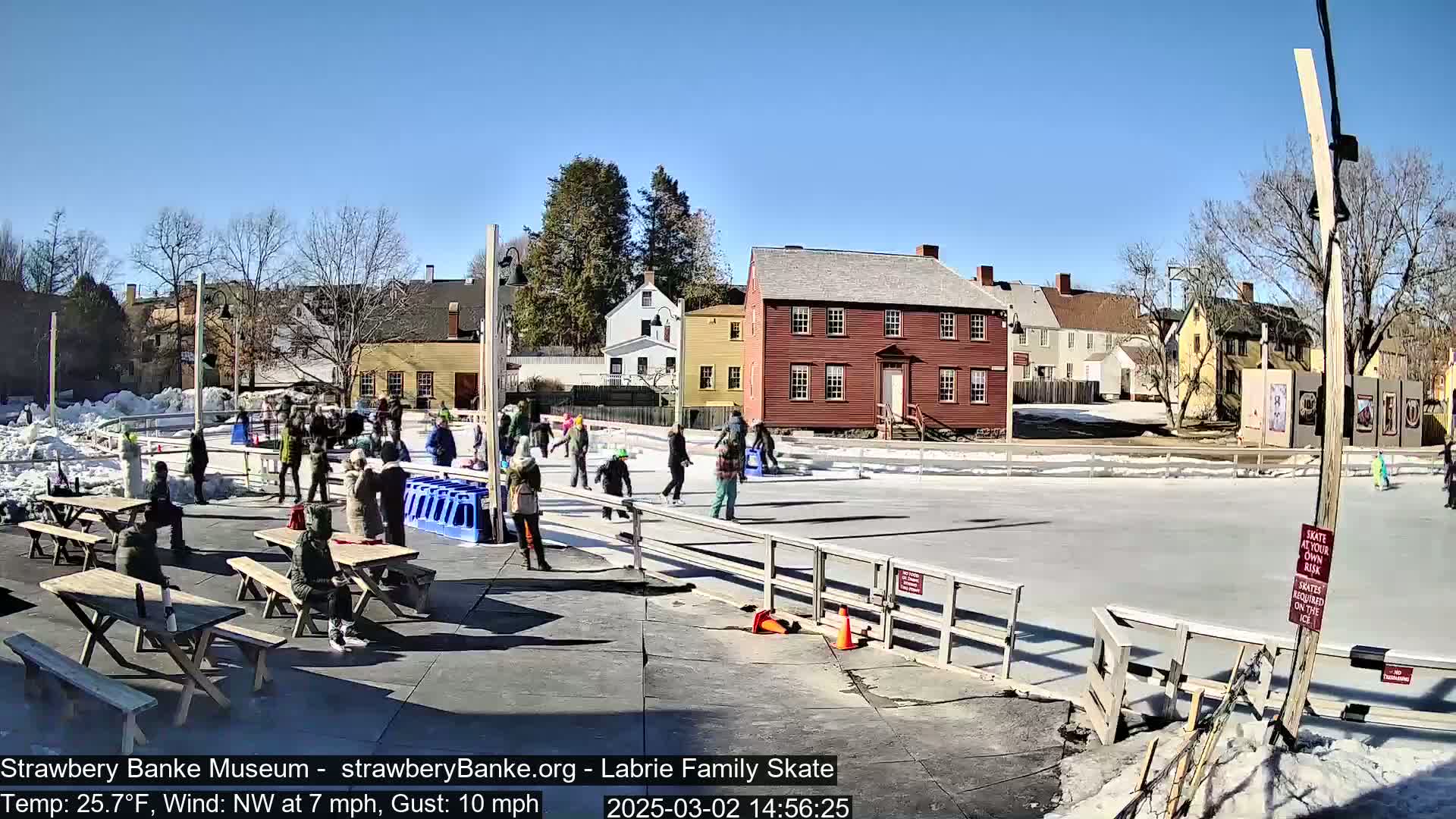 On a sunny day with a northwest wind, people ice skate on an outdoor rink surrounded by historic buildings and picnic tables.