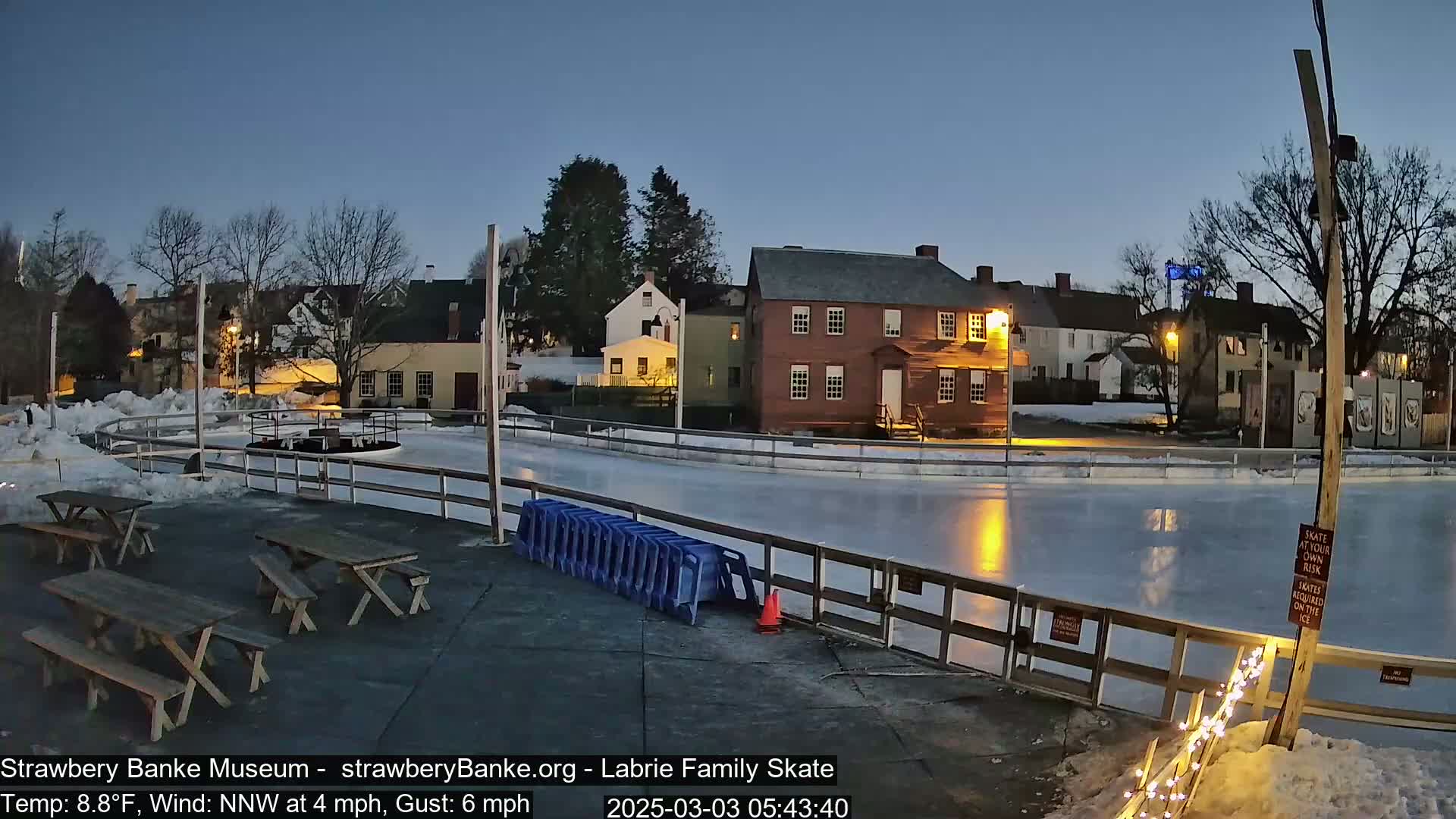 An outdoor ice rink at dusk, illuminated by lights, is surrounded by historic buildings and picnic tables, with a light snowfall on the ground.