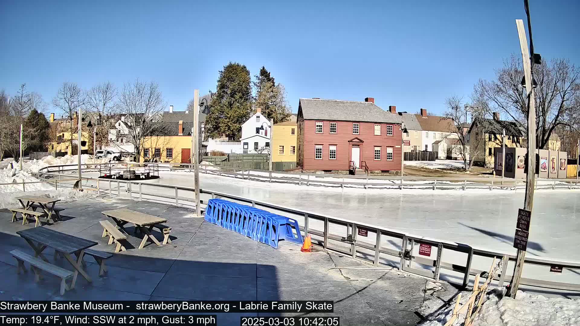 An outdoor ice skating rink, surrounded by historic-looking buildings and picnic tables, on a sunny day with a light south-southwesterly wind.