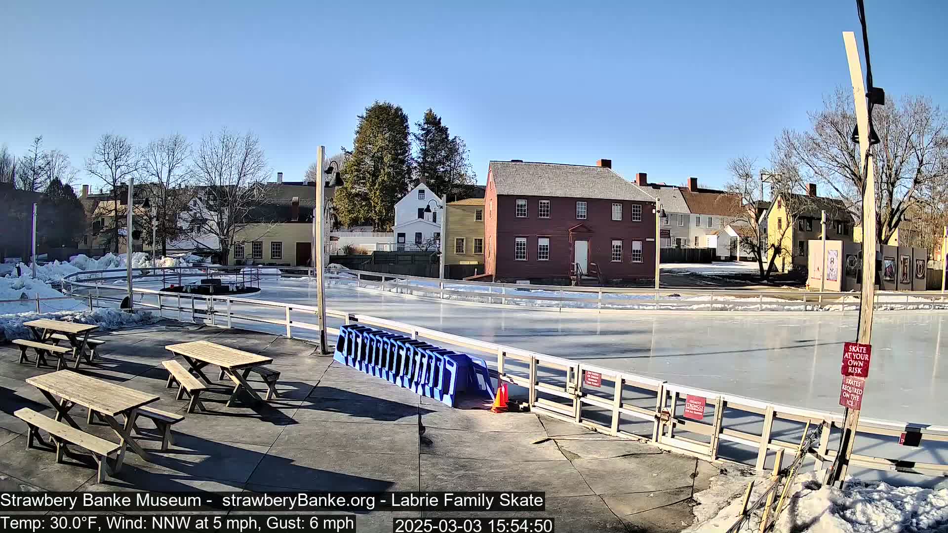 An outdoor ice rink surrounded by wooden picnic tables and historic buildings under a clear blue sky.